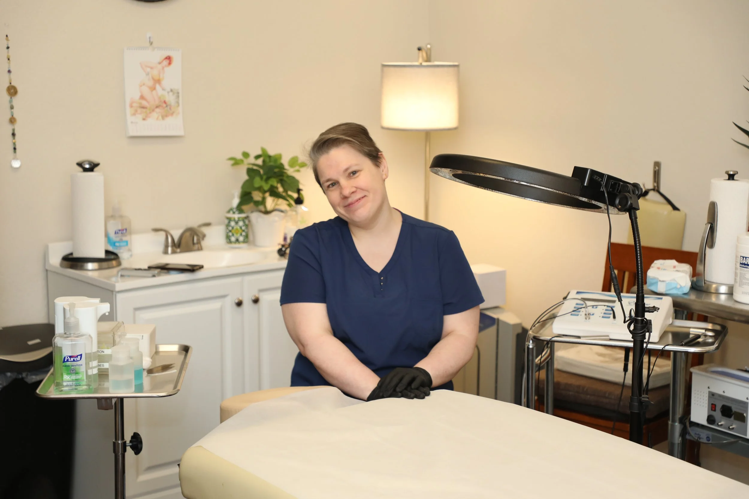 A woman with short hair wearing navy medical scrubs and black gloves sitting at a medical examination table in a clinic or medical room, smiling at the camera.