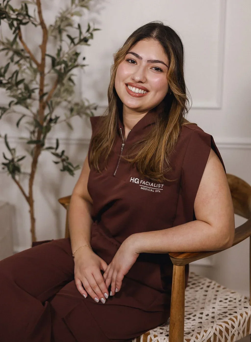 A woman with long brown hair and a warm smile sitting on a wooden chair in a professional uniform that says 'HG FACIALIST MEDICAL SPA'.