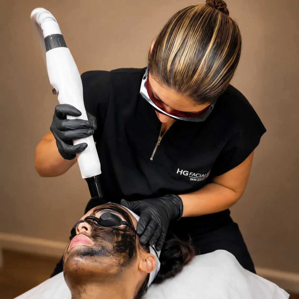 A woman receiving a facial treatment with a black mask, while a professional uses a specialized tool on her face in a spa or skincare clinic.