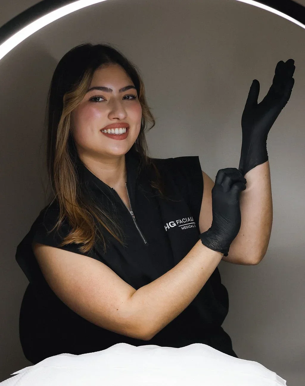 A woman with brown hair smiling and wearing black gloves, a black sleeveless top, and a black vest with a logo, standing inside a lighted circular frame.