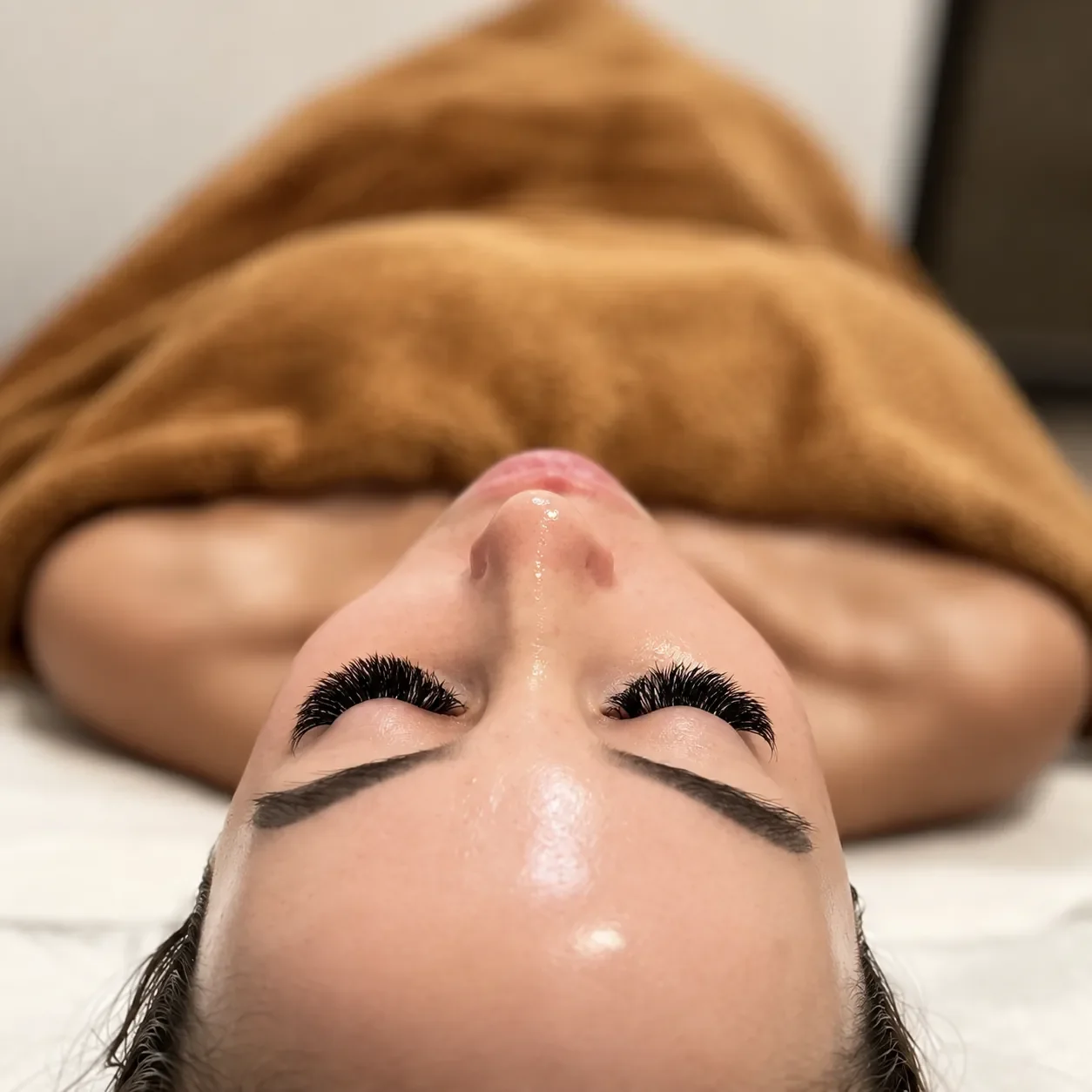 Close-up view of a woman lying down with her eyes closed, wearing thick eyelash extensions and a brown towel covering her body.