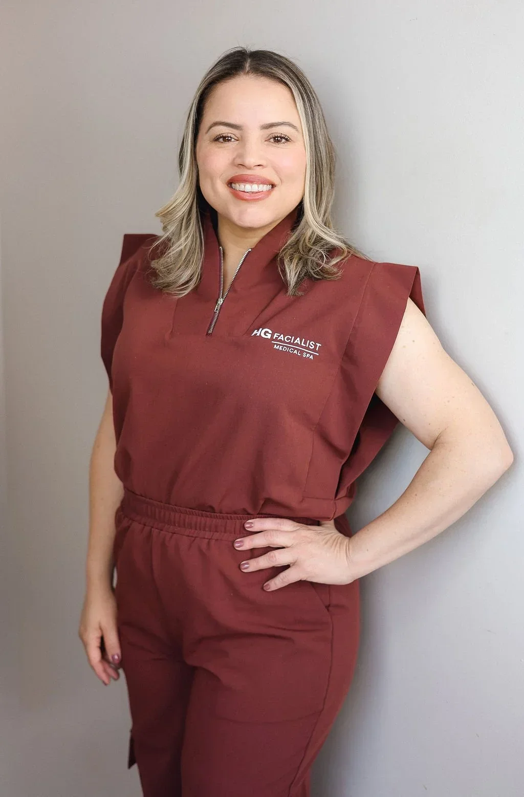 A woman with shoulder-length blonde hair, smiling, wearing a maroon medical spa uniform, standing against a plain light-colored wall.
