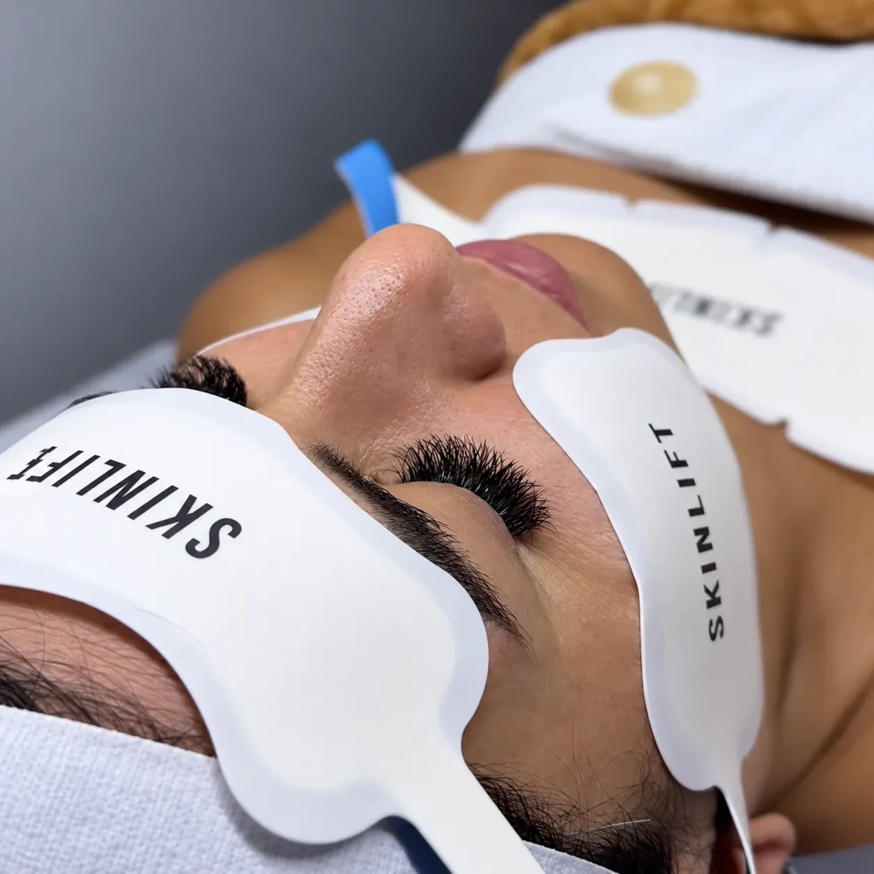 Close-up of a woman receiving a skincare or beauty treatment, lying on a bed with eye masks labeled 'SKINLIFT' over her closed eyes, with eyelash extensions visible.