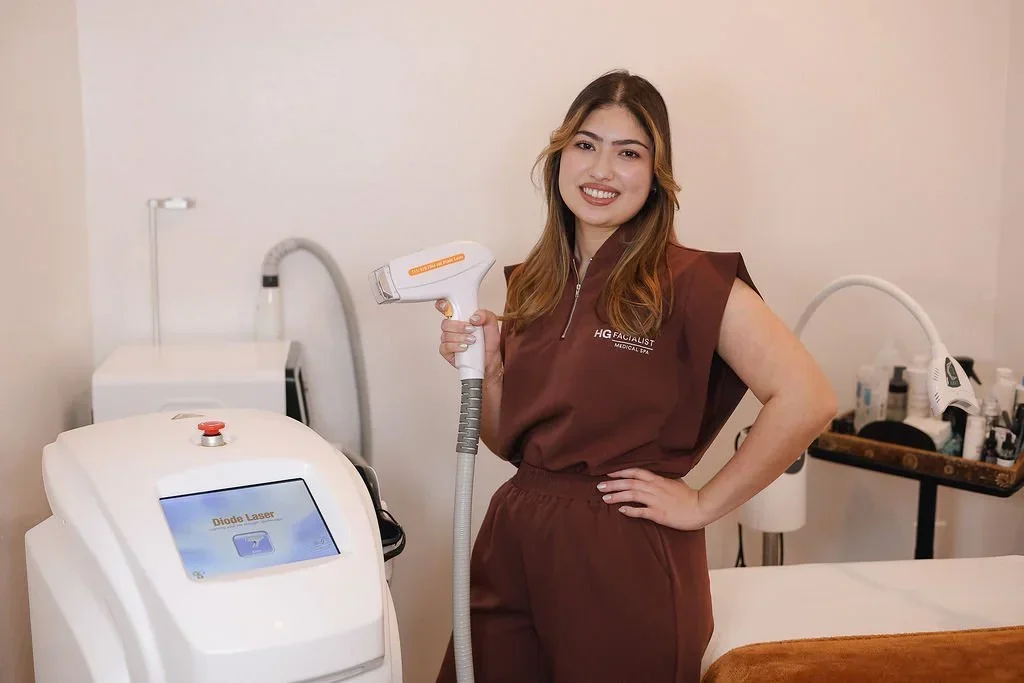 A woman with light brown hair holding a laser device in a clinic room, smiling, standing next to a laser machine labeled 'Diode Laser'.