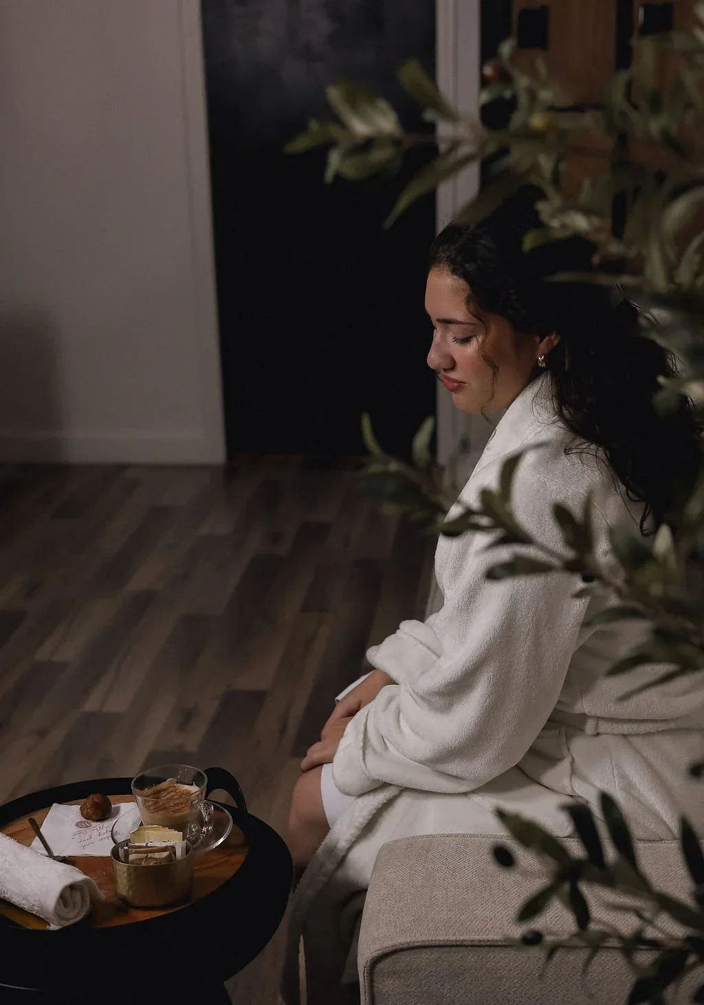 A woman with curly dark hair, wearing a white robe, sitting on a beige couch with her hands on her lap, in a cozy room with wooden flooring and a dark background, with a small round table nearby holding a glass of tea or coffee, a plate with a snack, and some packaged foods.