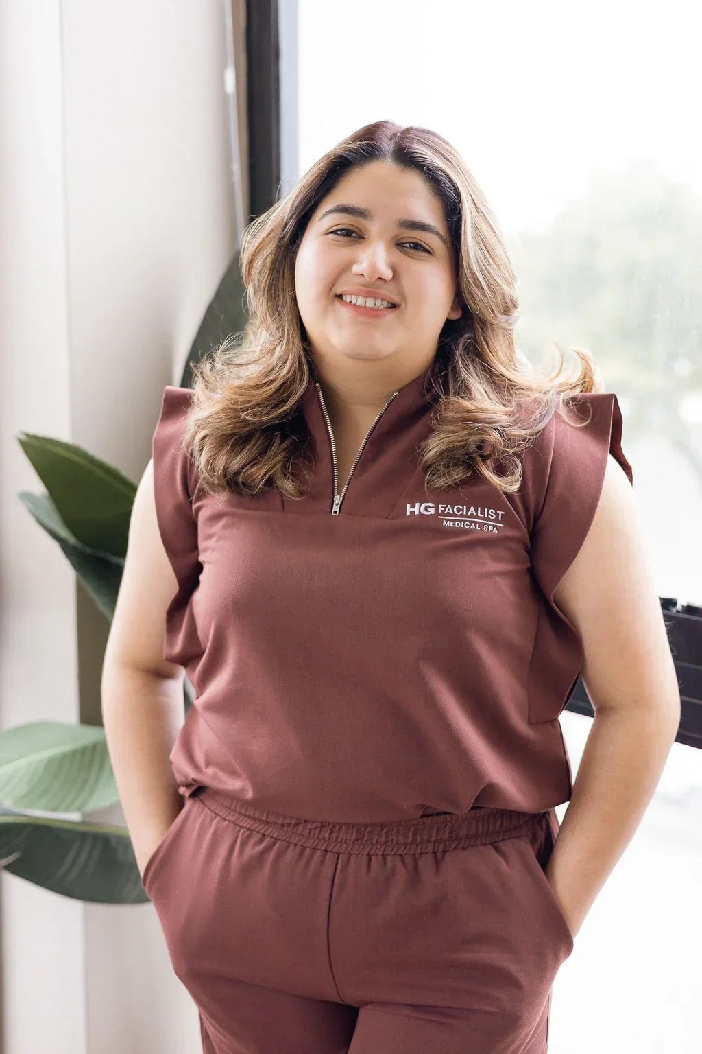 A woman with shoulder-length wavy hair smiling, wearing a brown sleeveless outfit with a zipper and the logo 'HG FACIALIST' on it, standing indoors near a window and a large green plant.