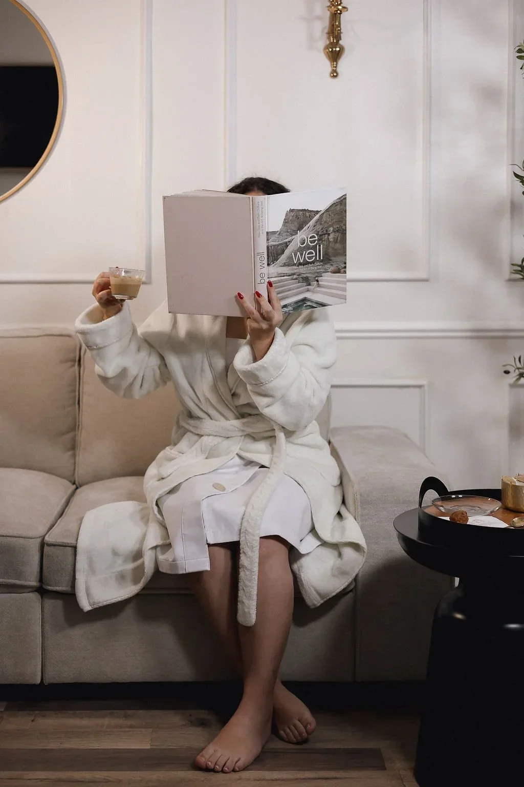 A woman sitting on a beige sofa, wearing a white bathrobe and a white knee-length dress, holding a cup of coffee in one hand and a book titled 'be well' in front of her face. The room has white walls with decorative paneling, a round mirror on the left, a small black side table to the right with a tray of food, and a plant on the far right.