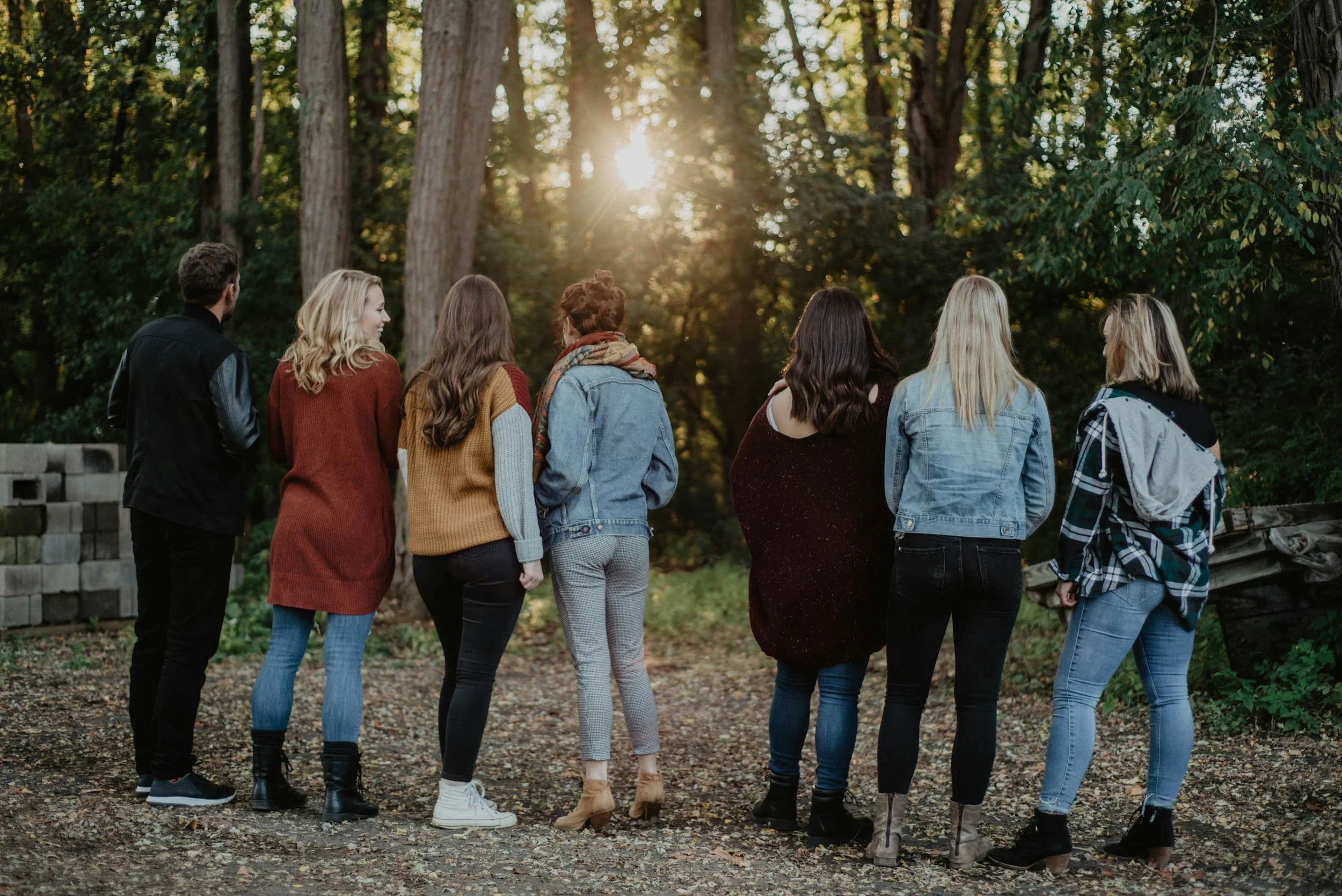 Group of seven young women standing outdoors in a forest, facing away from the camera, with sunlight filtering through trees.