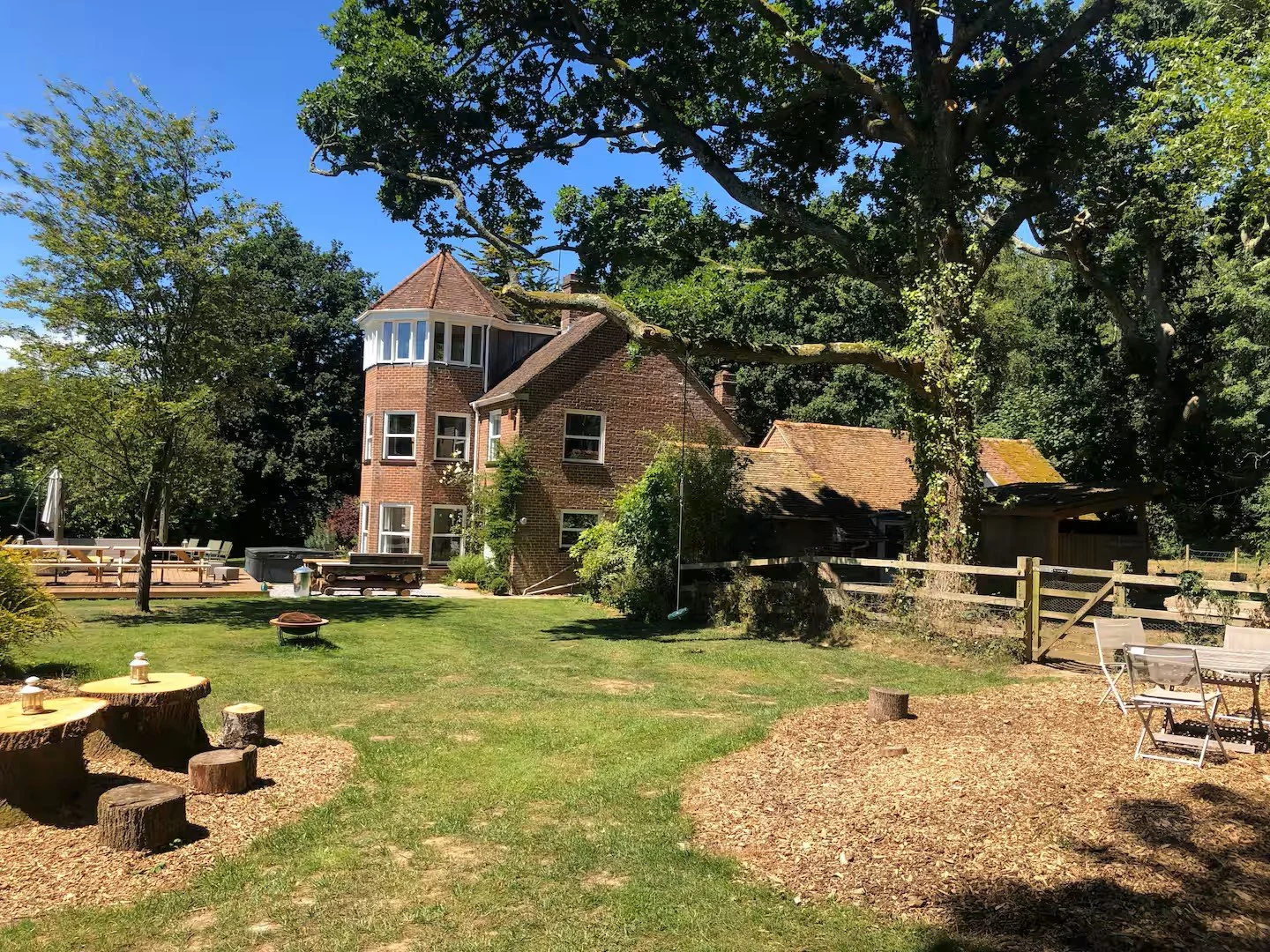 Large garden with green grass, mature trees, and outdoor seating area including tables and chairs. A multi-story brick house with a turret and a separate garage are visible in the background, along with a wooden fence and a swing hanging from a tree.