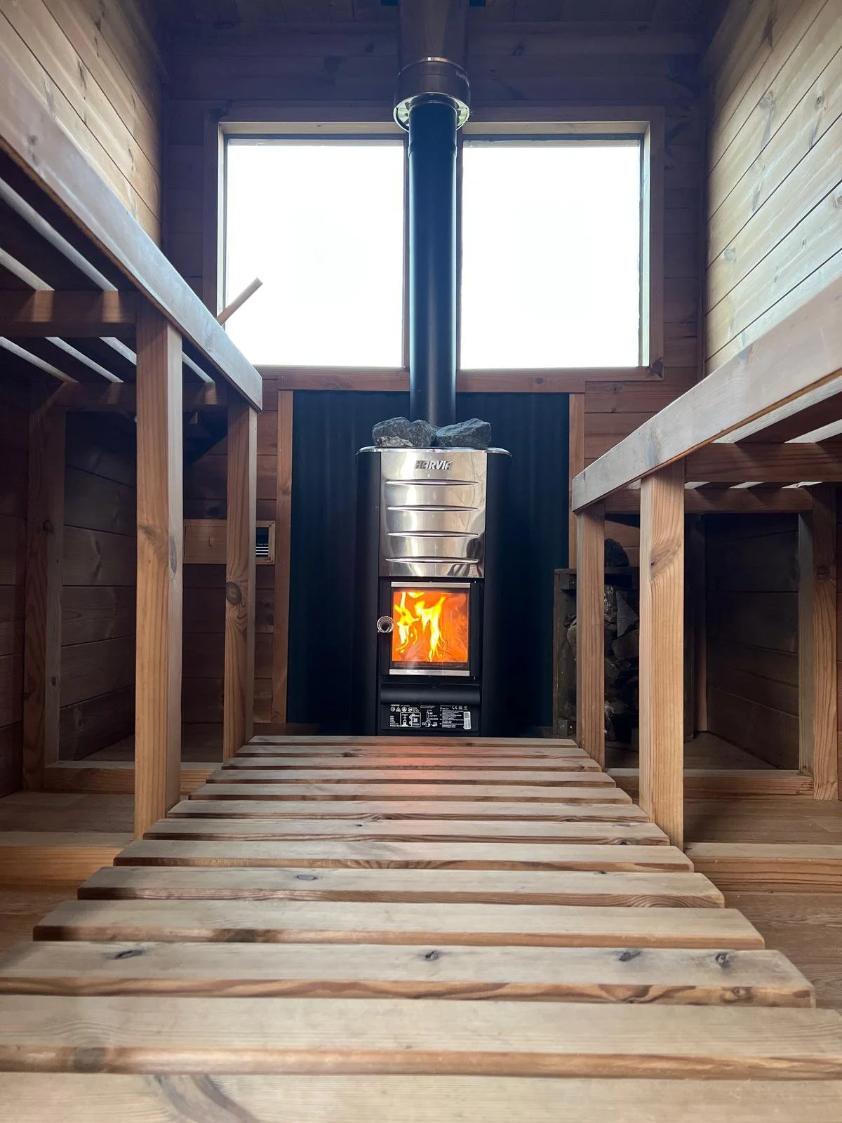 A wood-burning stove with a fire inside, situated in a sauna with wood walls and built-in wooden benches on each side.