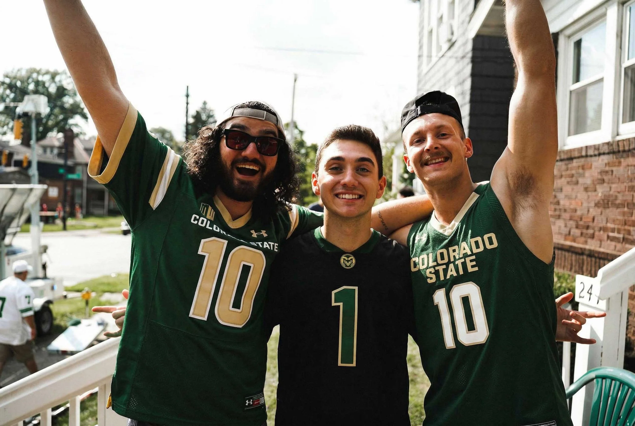 Three young men wearing Colorado State football jerseys smiling and celebrating outdoors on a sunny day, standing on a porch with a street and house in the background.