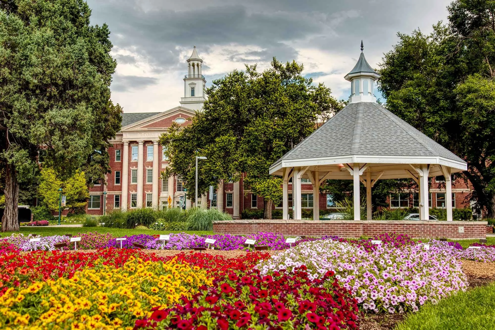 A park with colorful flowers, a white gazebo, tall trees, and a red brick building with white columns in the background.