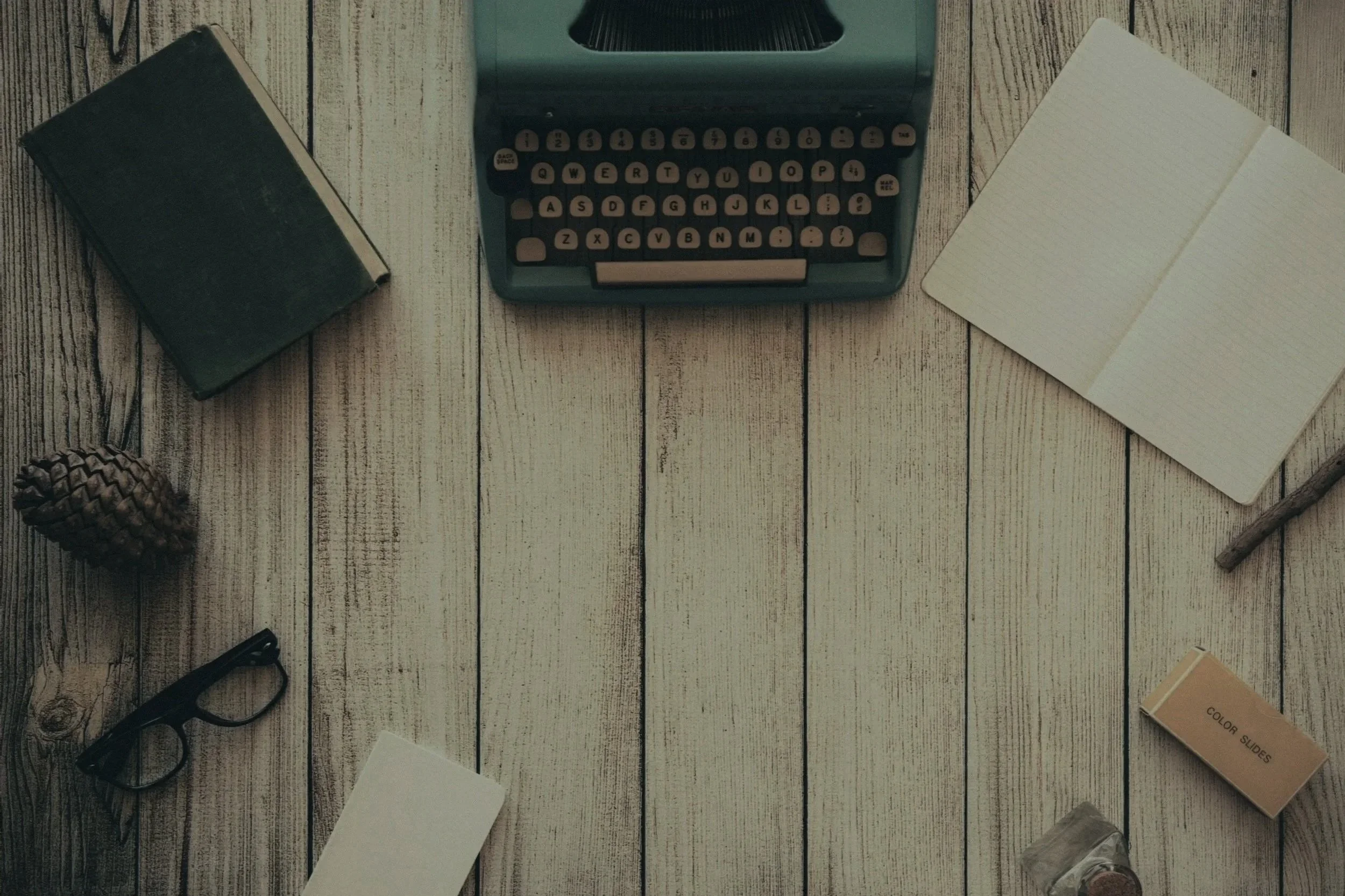 Flat lay image of vintage typewriter, closed notebook, open notepad, pine cone, glasses, box of color slides, and small jar on a wooden surface.