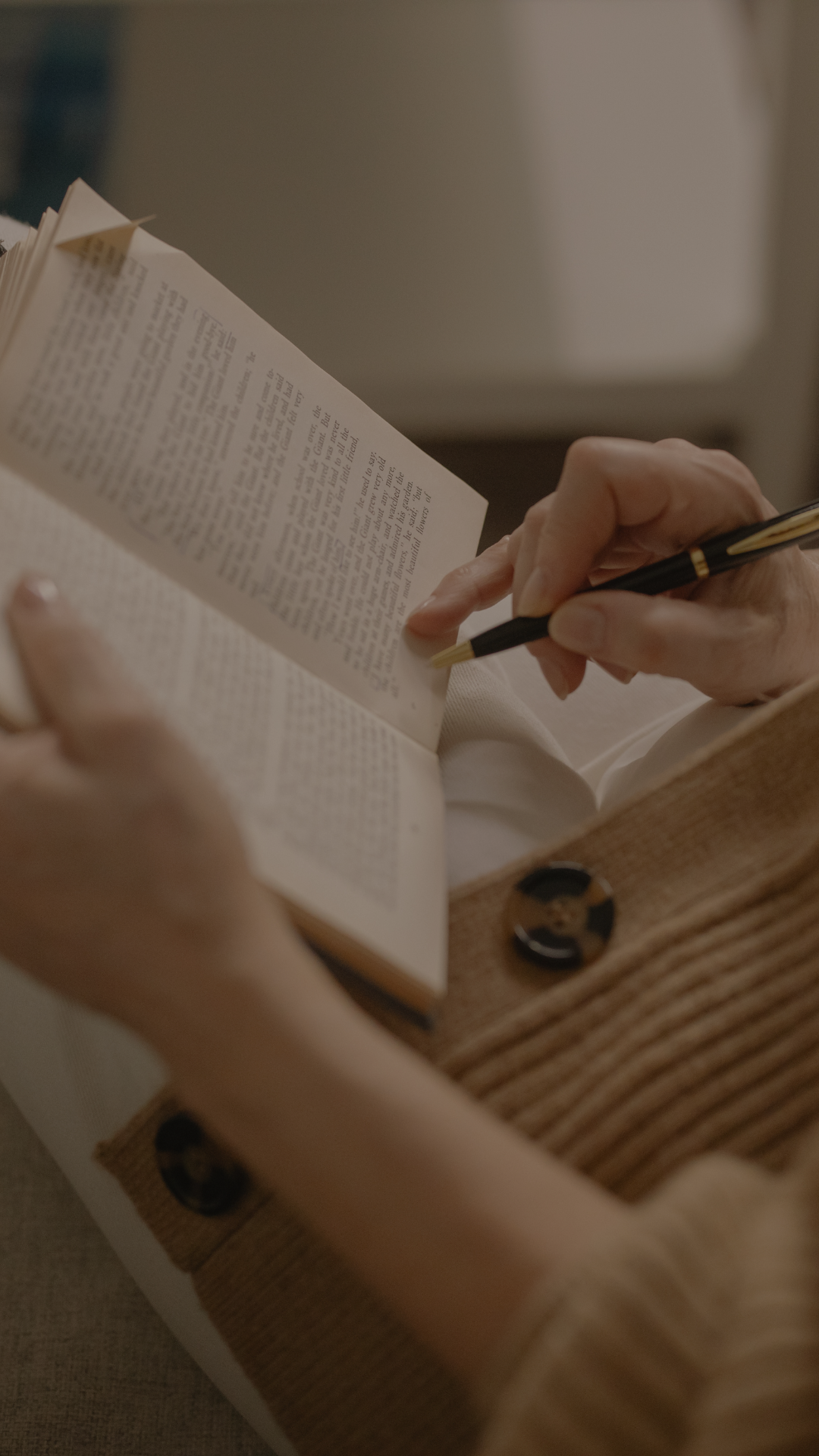 A person holding a pen and a book, writing or taking notes at a wooden desk in a softly lit setting.