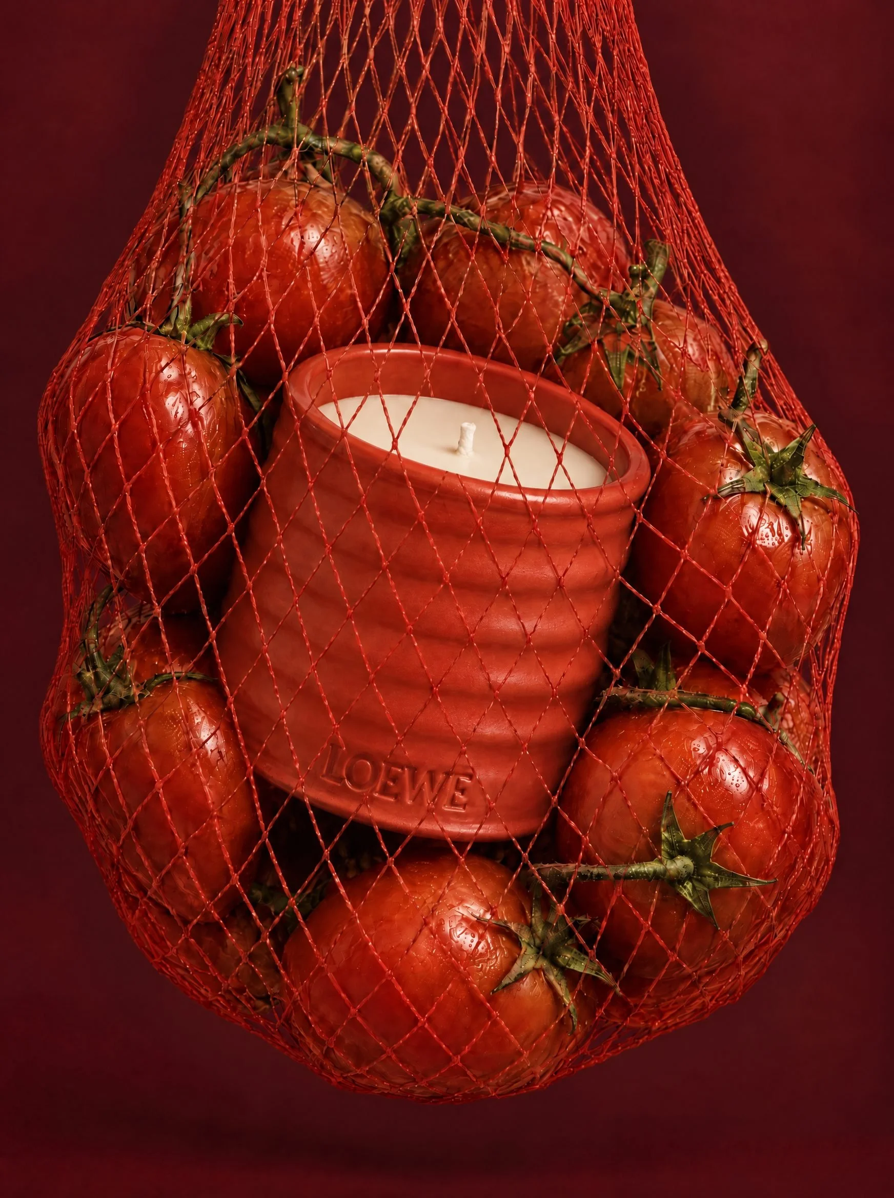 The red ceramic LOEWE candle held in a red net bag of ripe vine tomatoes