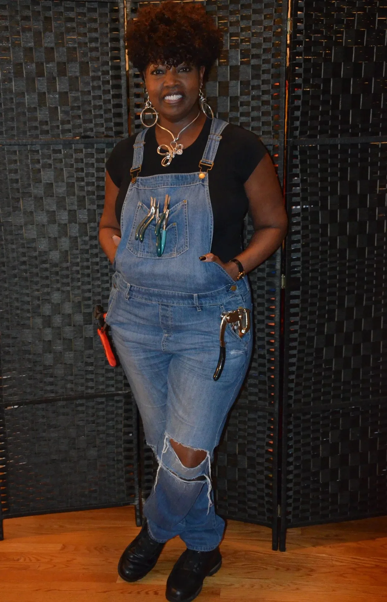 A woman with dark skin and curly hair wearing a black t-shirt, denim overalls with tools in the pocket, ripped jeans, and black boots, standing against a black decorative screen and wooden floor.