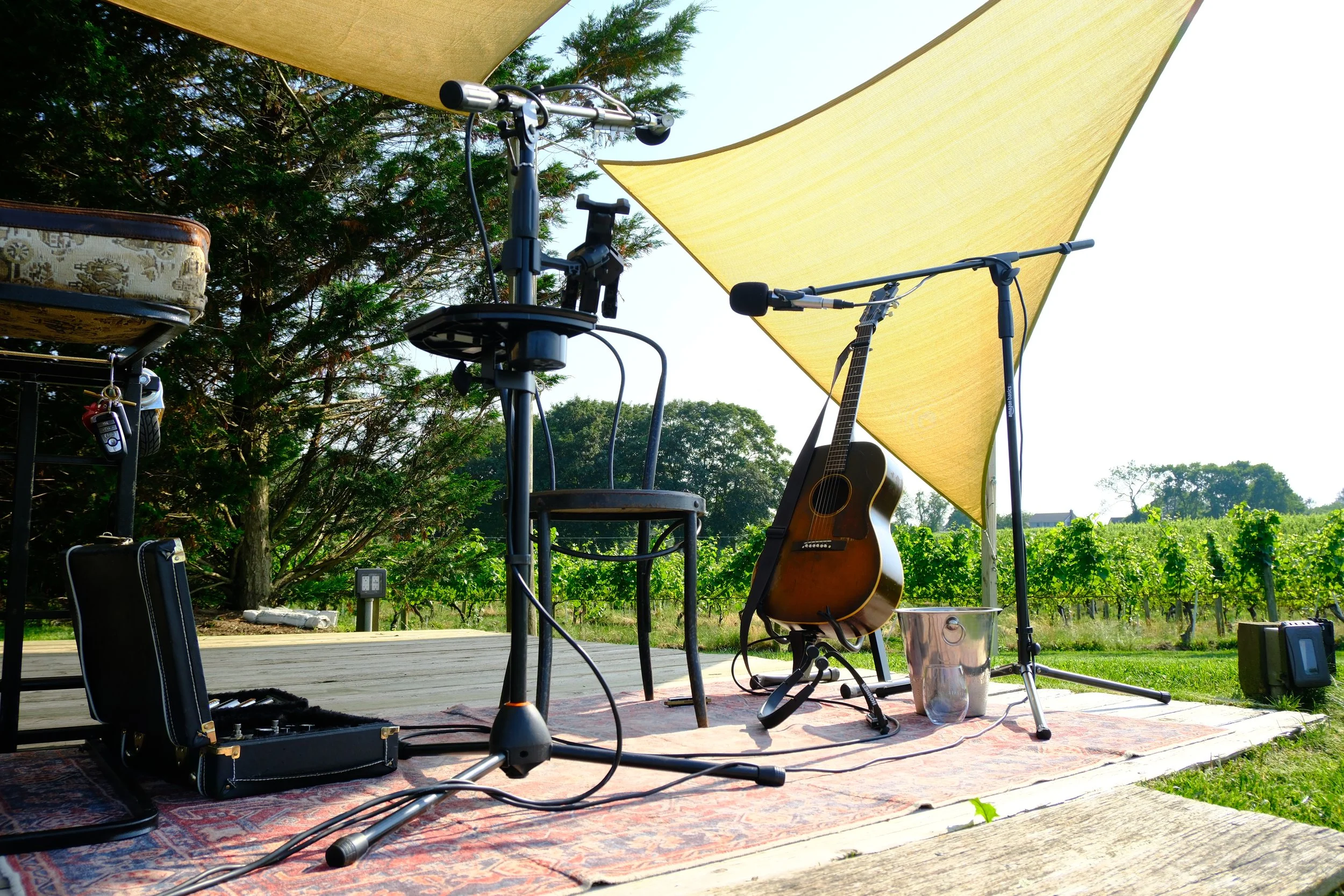 Musical setup outdoors on a wooden stage with a guitar, microphone, and amplifier under yellow shade sails, surrounded by greenery and vines in the background.