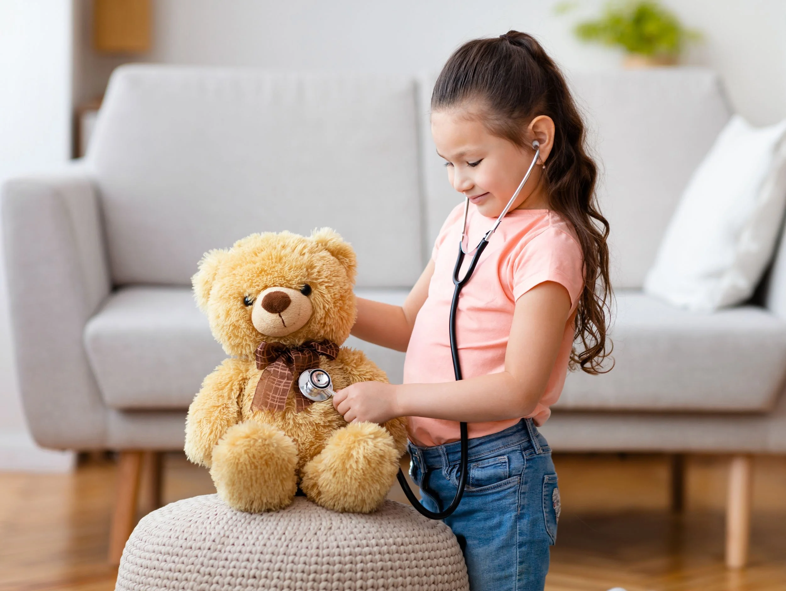 Young girl in pink shirt and jeans with stethoscope listening to teddy bear's chest at home.