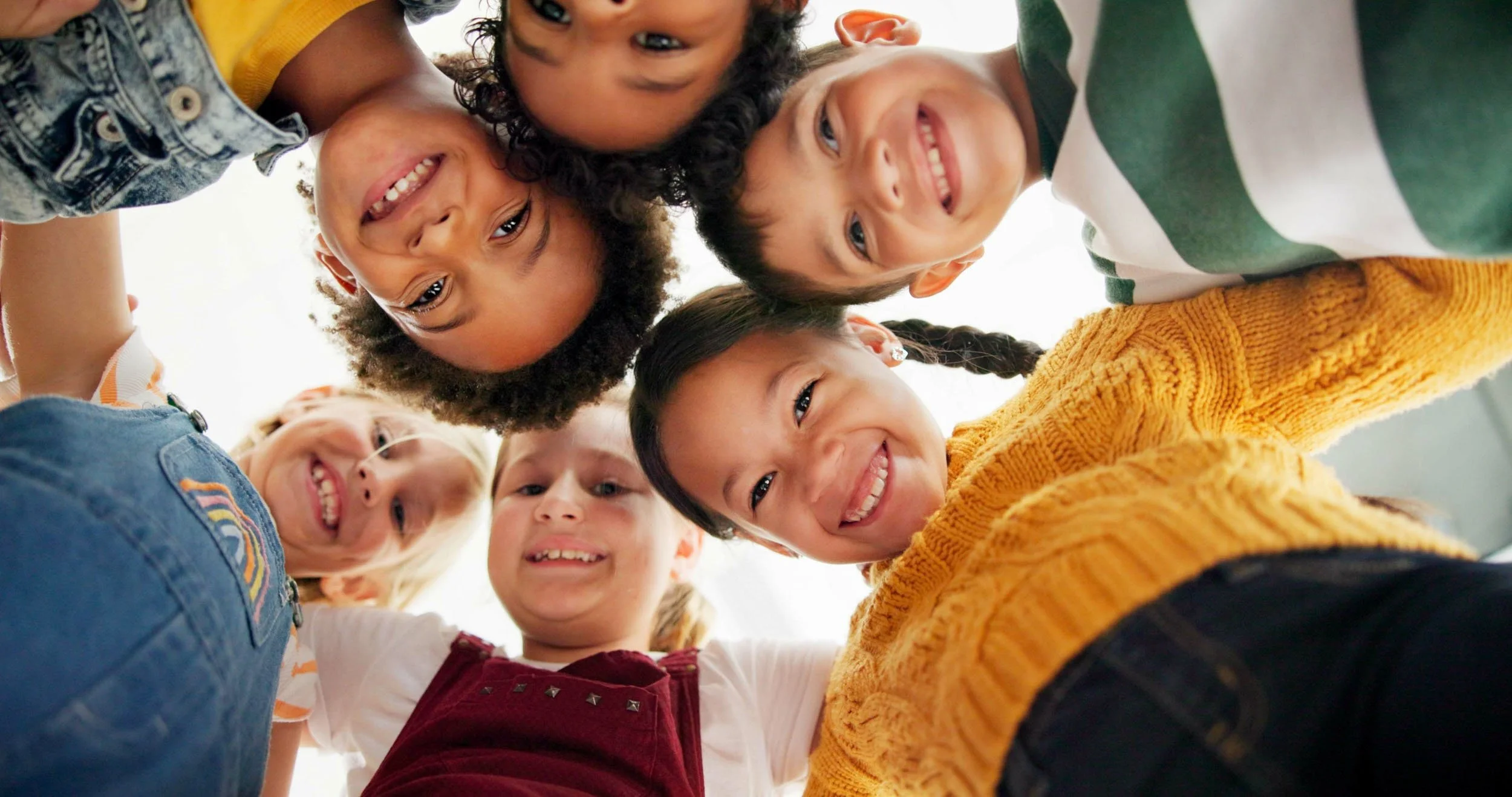 Group of children in a huddle, smiling and looking down into the camera, taken from below.