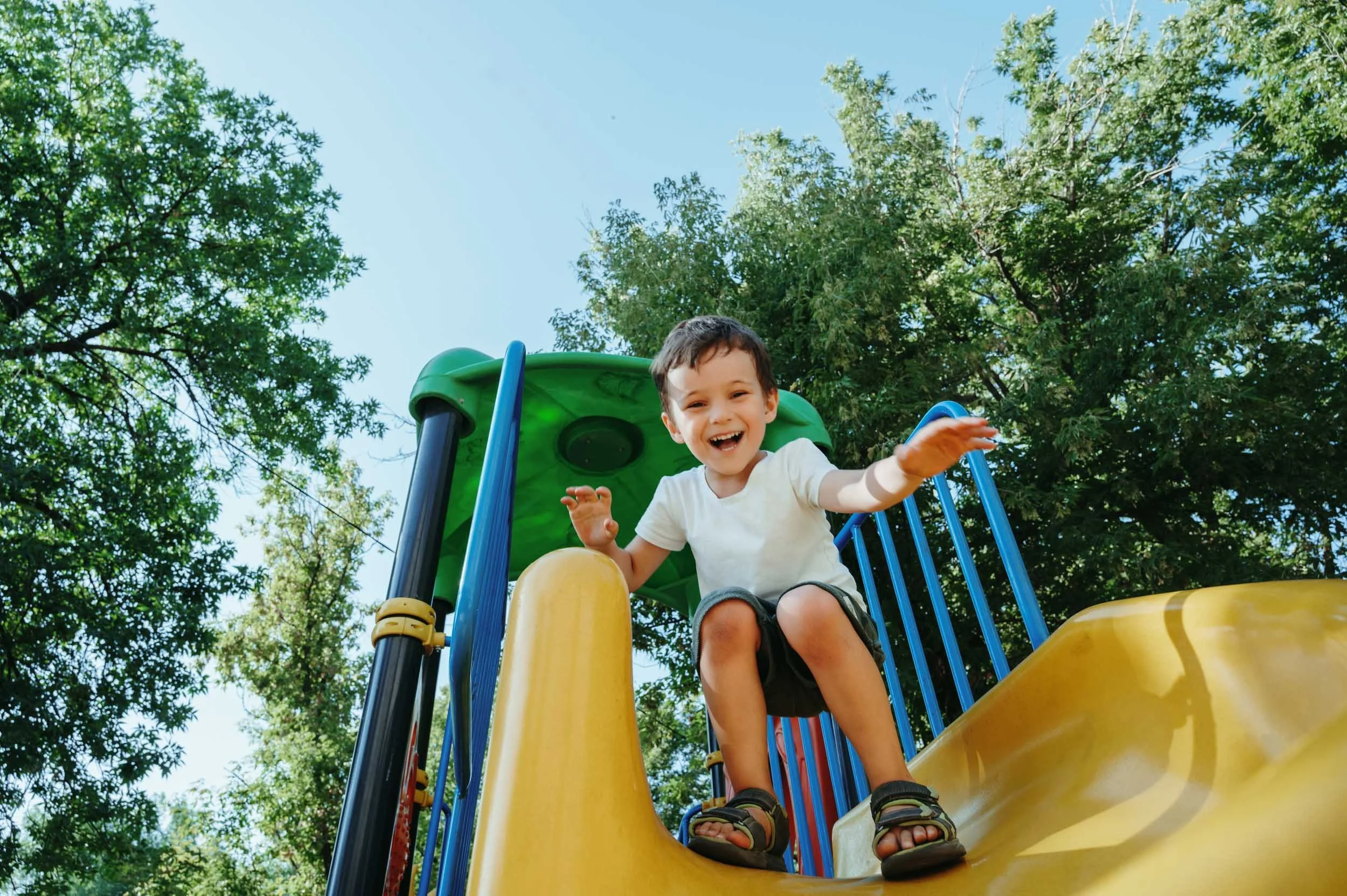 A young boy with short dark hair and a big smile sitting at the top of a yellow slide on a playground, surrounded by green trees and blue sky.
