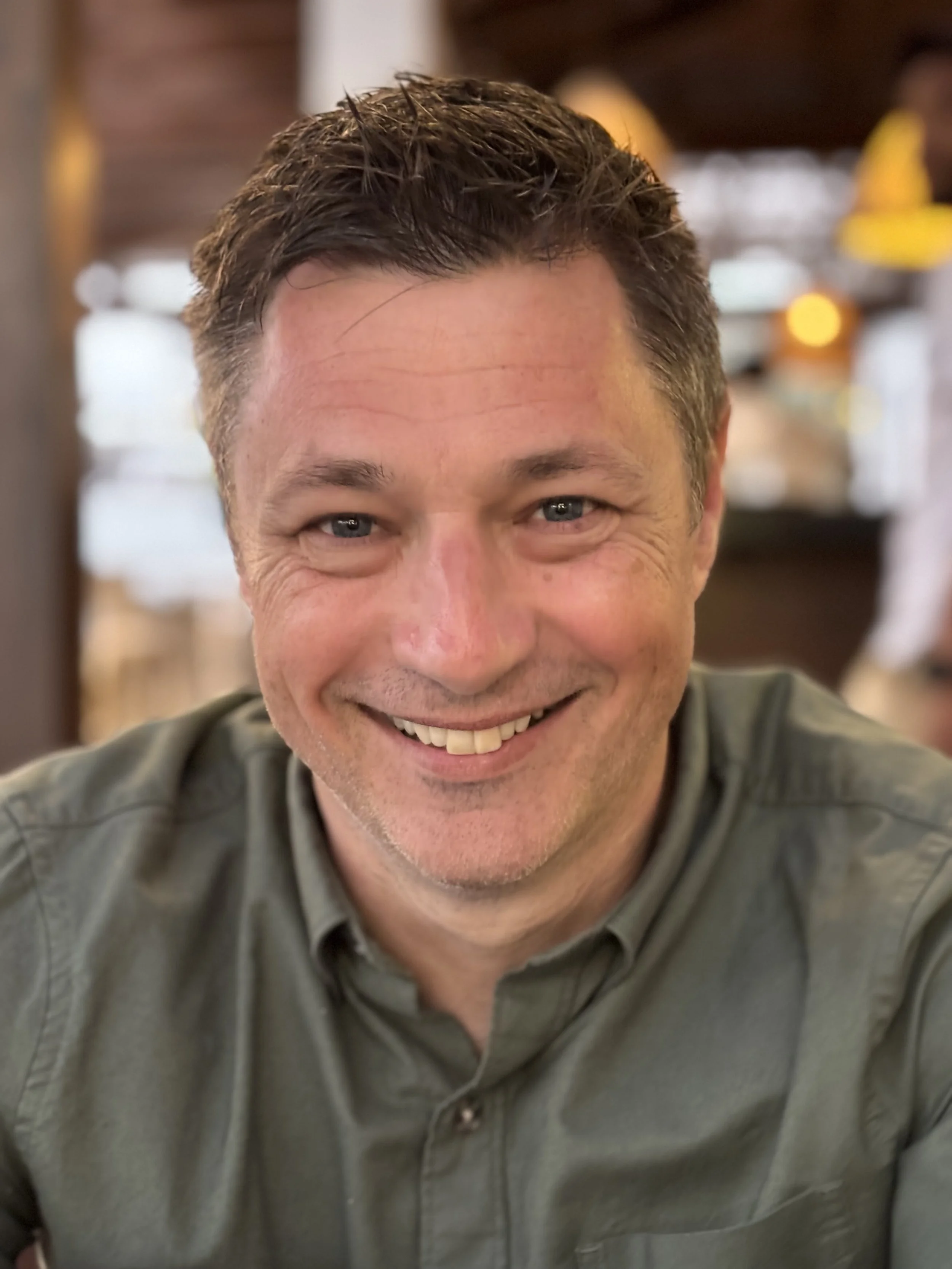 A man smiling at a camera in a cozy, warmly lit setting, possibly a restaurant or café.