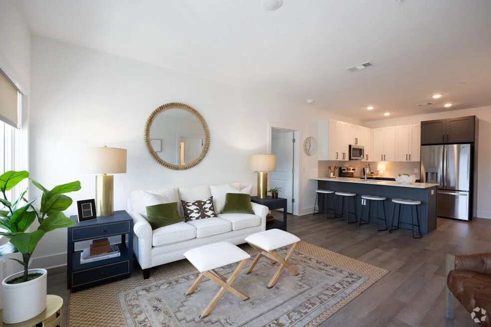Living room with white sofa, wooden stools, and a kitchen with gray cabinetry and stainless steel appliances. One Bedroom apartment in Gainesville Georgia.