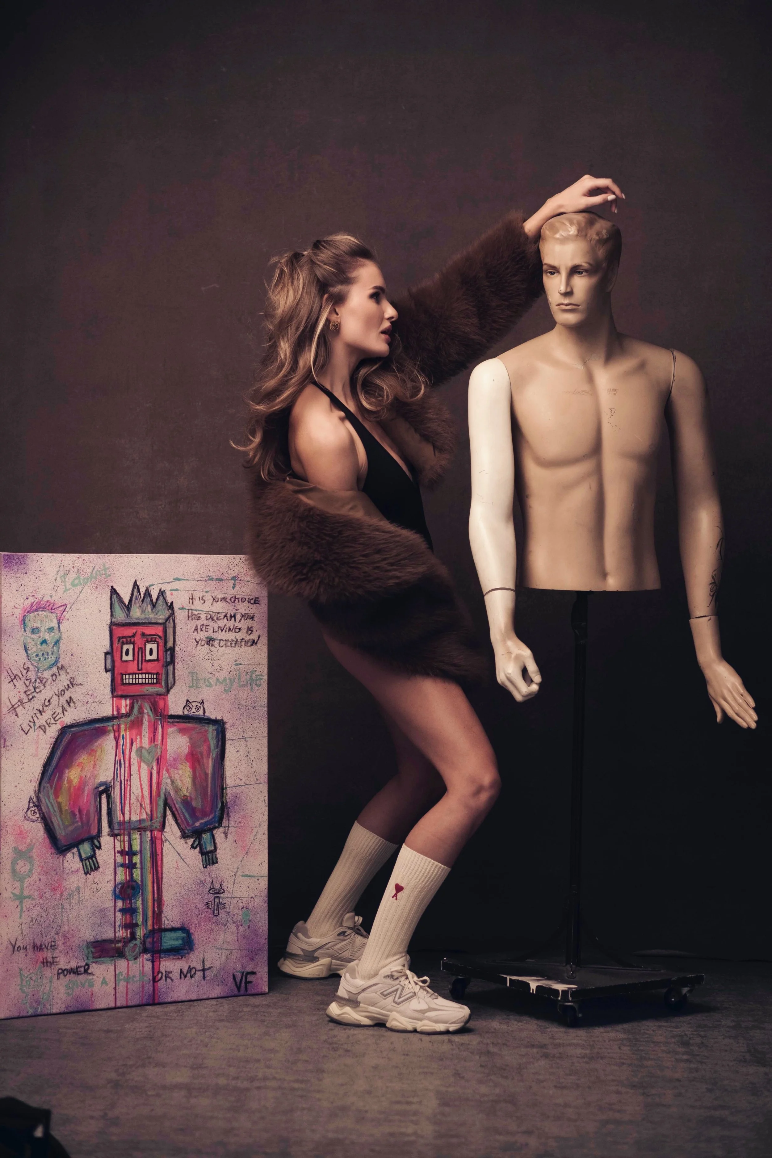 A woman wearing a brown fur coat, black dress, white sneakers, and cream knee-high socks with a red ribbon stands next to a mannequin. The woman is touching the mannequin's head. There is a colorful drawing on the floor depicting a robot-like figure with graffiti and text around it.