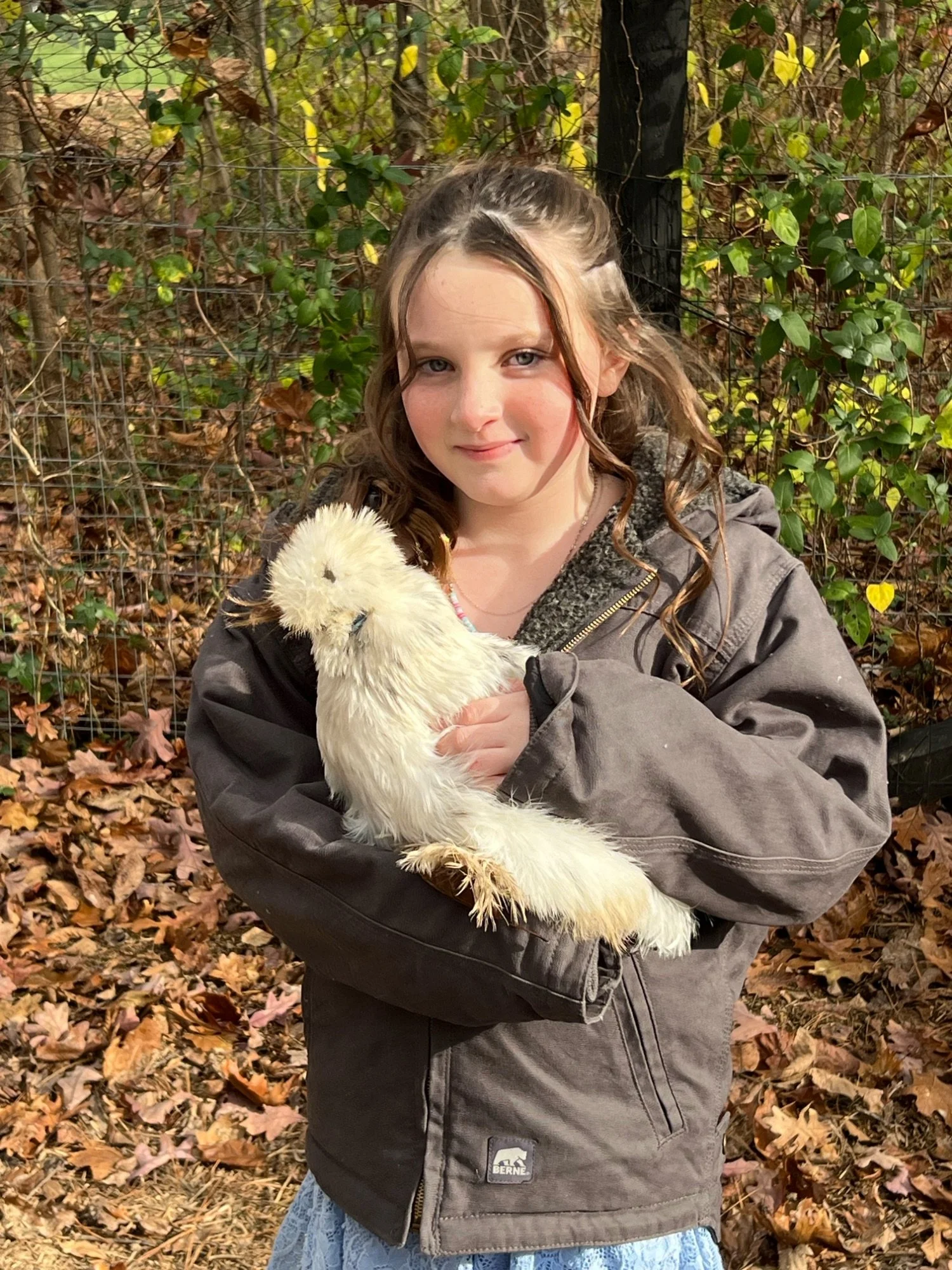 A young girl with long, curly hair and light skin is holding a fluffy white duckling and standing outdoors on a fall day with fallen leaves on the ground and green and yellow leaves on the bushes behind her.