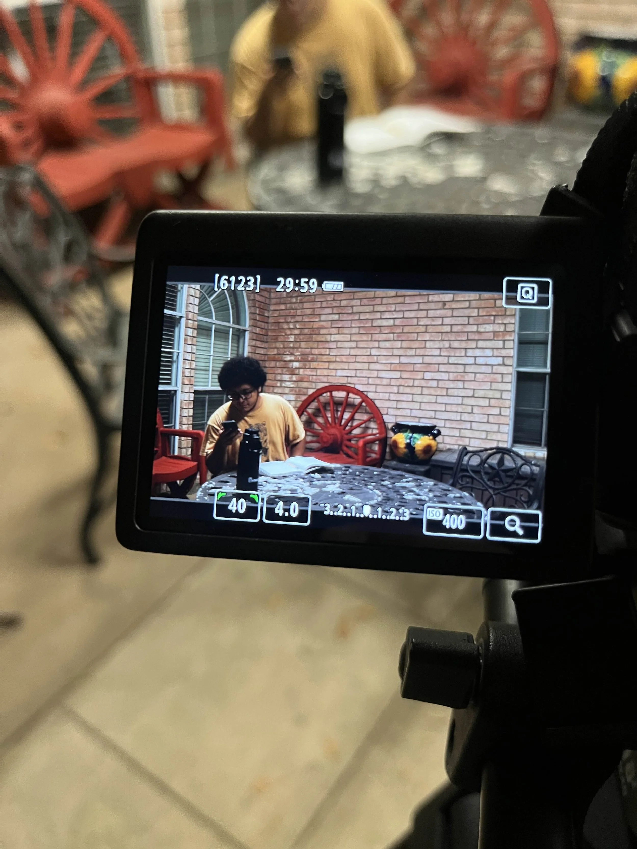 Photo of a person with curly black hair wearing glasses and a yellow shirt, looking at a phone, sitting at an outdoor table with a brick wall background and red chairs, taken through a camera viewfinder.