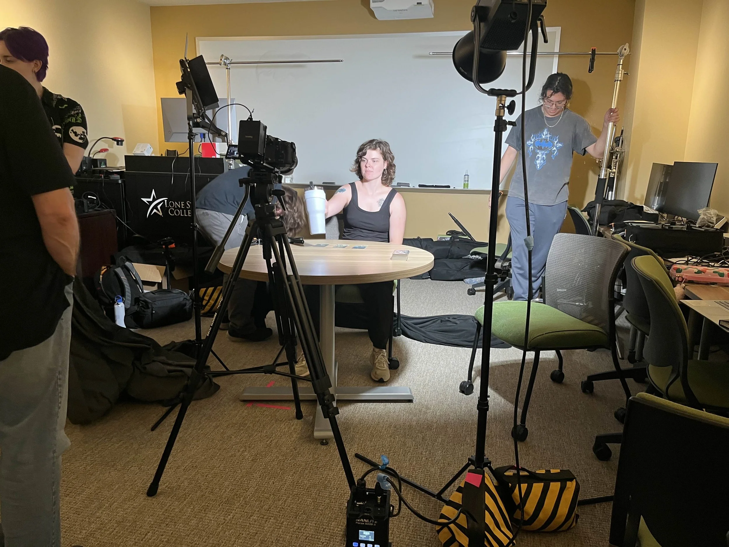 A woman with curly hair wearing a black tank top reflects on her face while sitting at a table in a classroom with a whiteboard behind her. People are involved in a filming or photography setup with cameras, lights, and equipment around her.