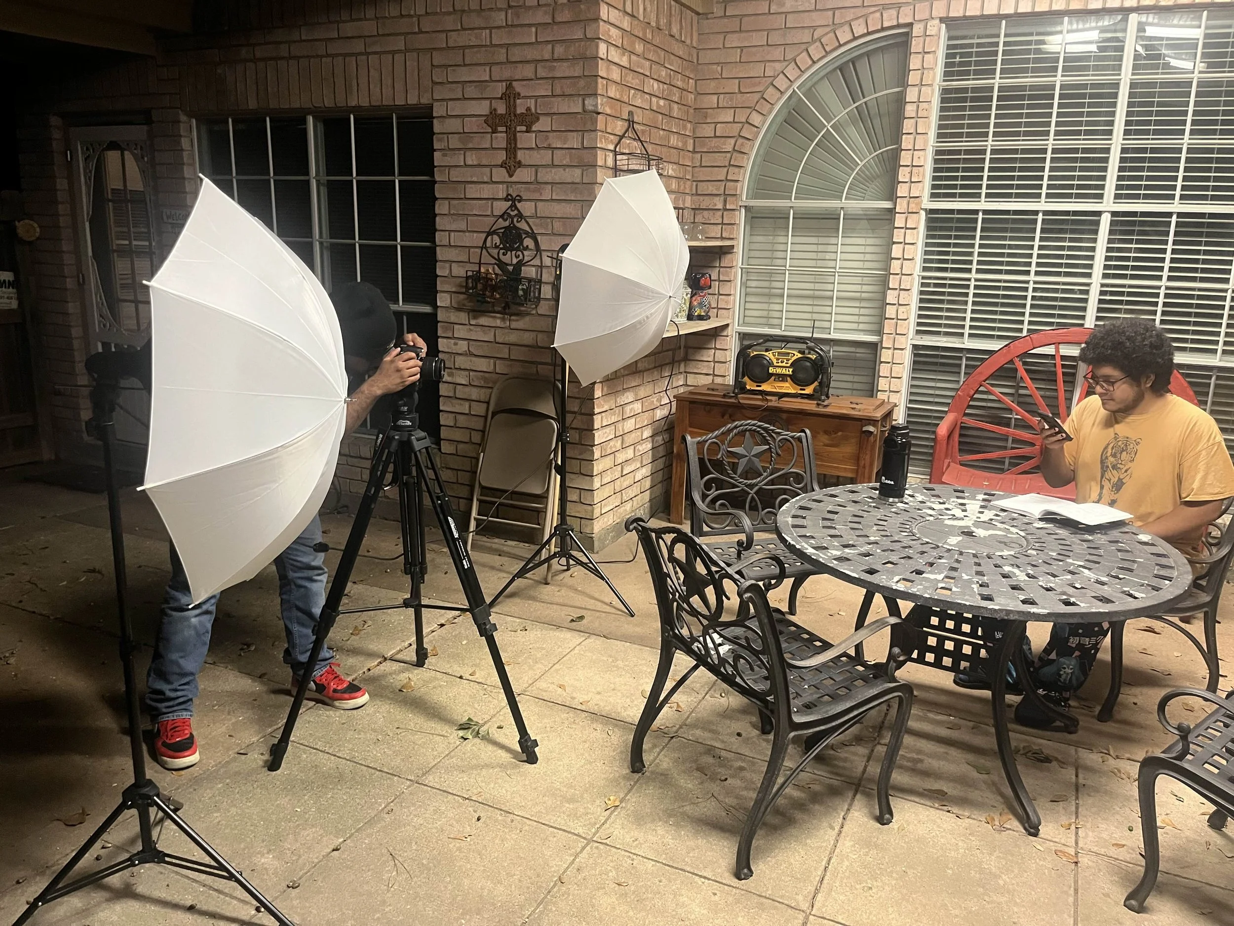 A patio scene with two large white umbrellas set up, one in use by a person taking a photo with a camera, and the other in front of a man sitting at a table using a smartphone. There is outdoor furniture, a brick wall with a cross and decorative items, and large windows with blinds in the background.