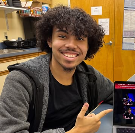A young man with curly hair and a beard smiling while sitting at a table, pointing to a laptop, in a room with wooden walls and shelves.