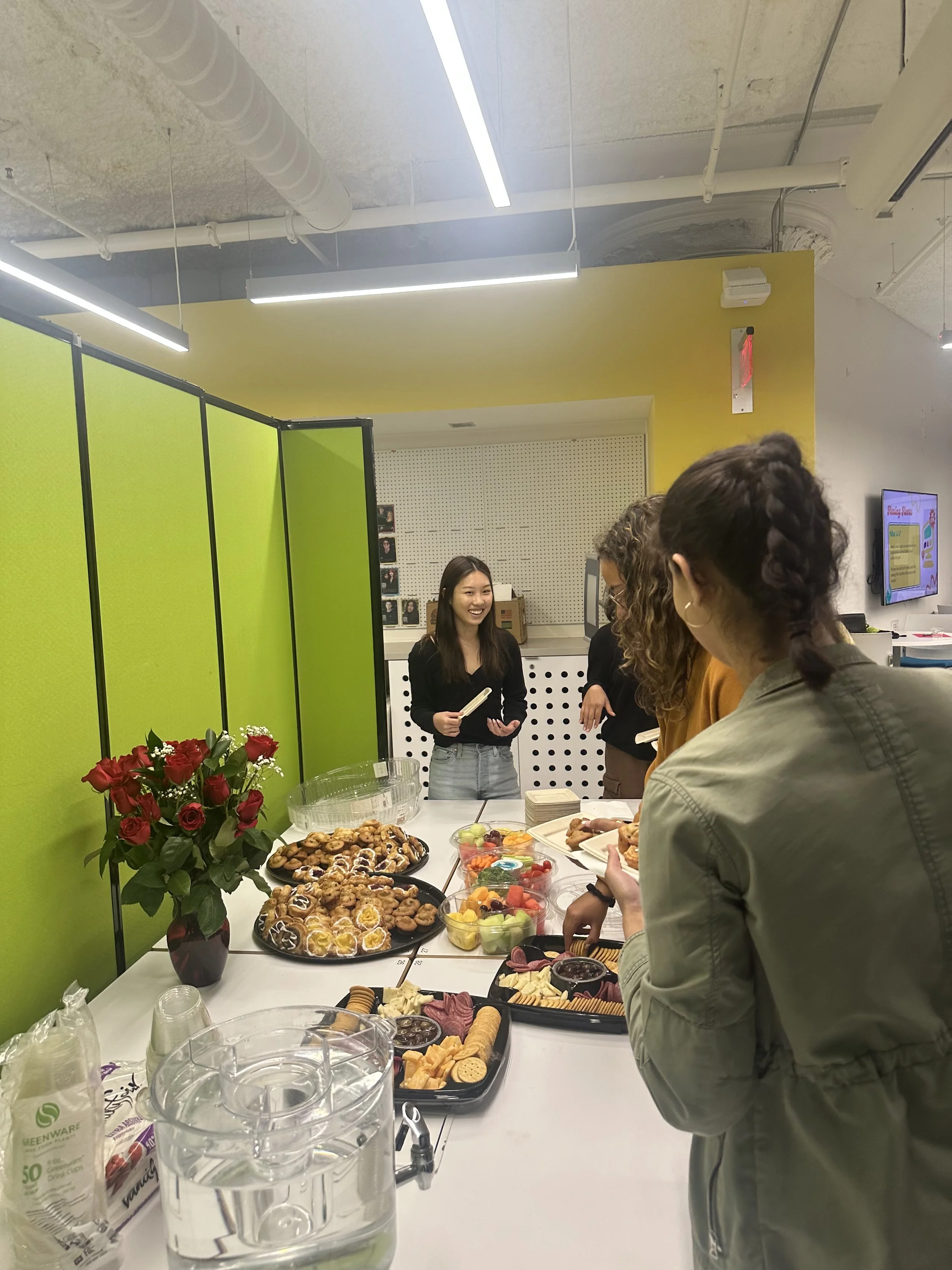 People gathered around a table with assorted snacks and desserts, with a woman smiling in the background in an office setting.