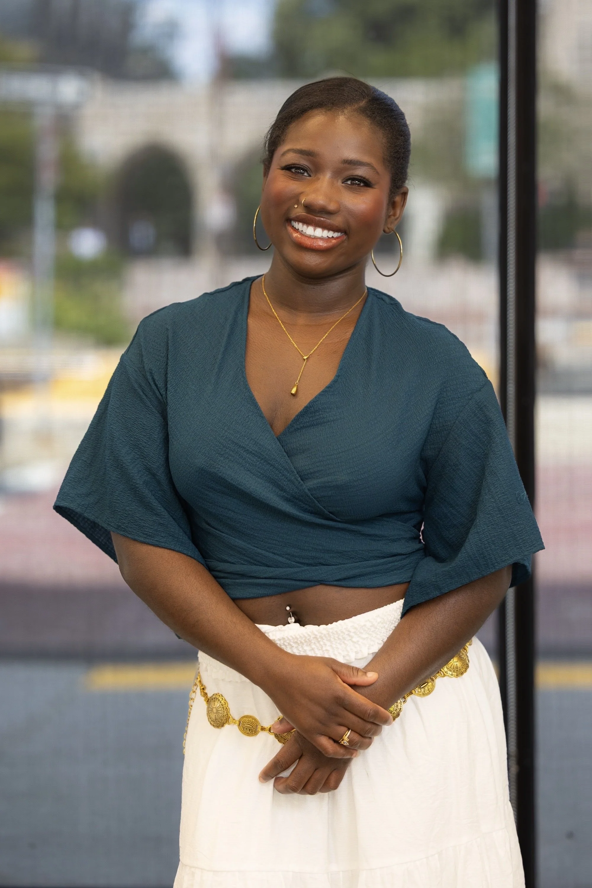 Smiling woman with dark hair, wearing a teal wrap top, white skirt with gold chain belt, and gold jewelry, standing indoors in front of a window.