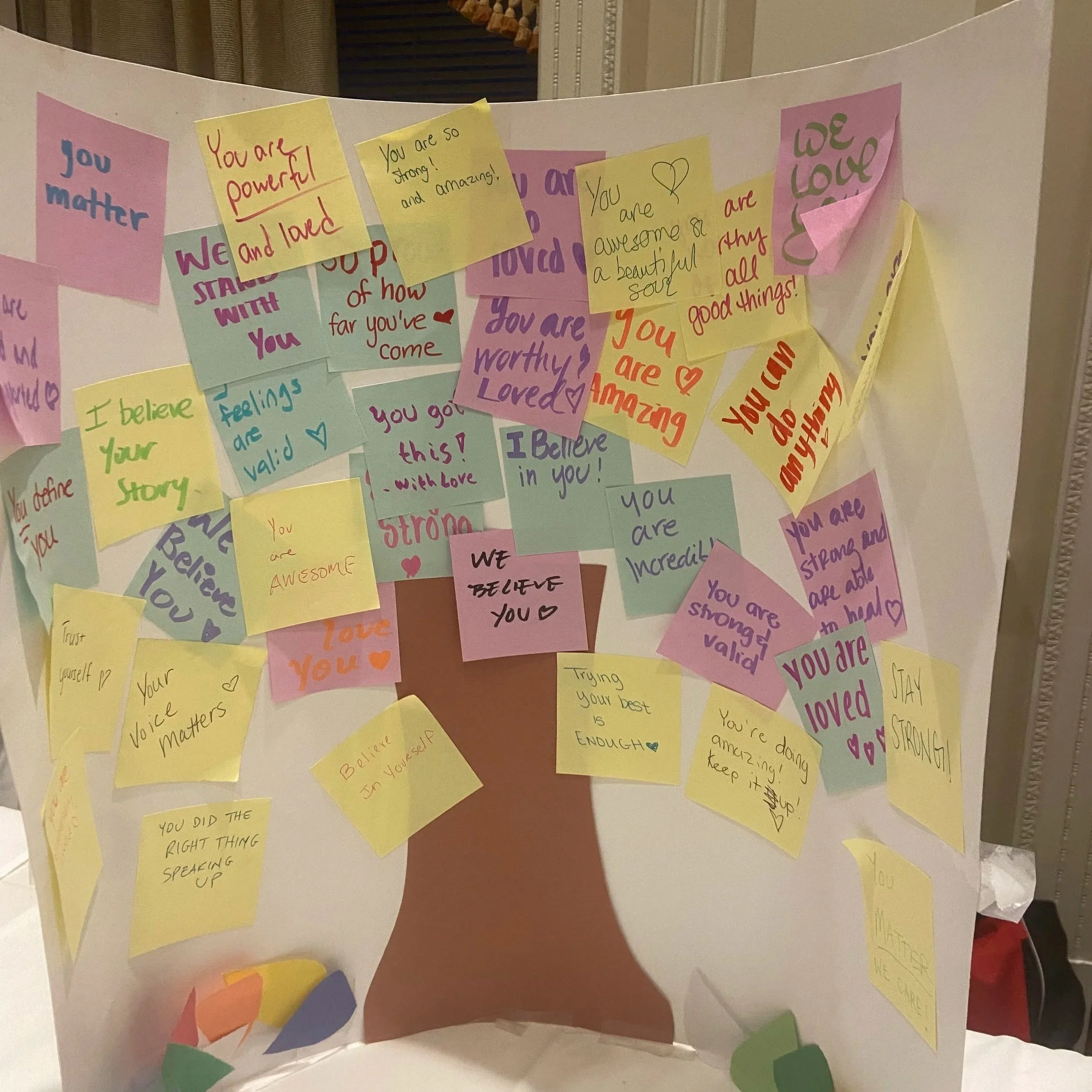 A colorful display board with numerous hand-written notes and messages of encouragement and love, featuring a brown paper tree trunk and various multicolored leaves.