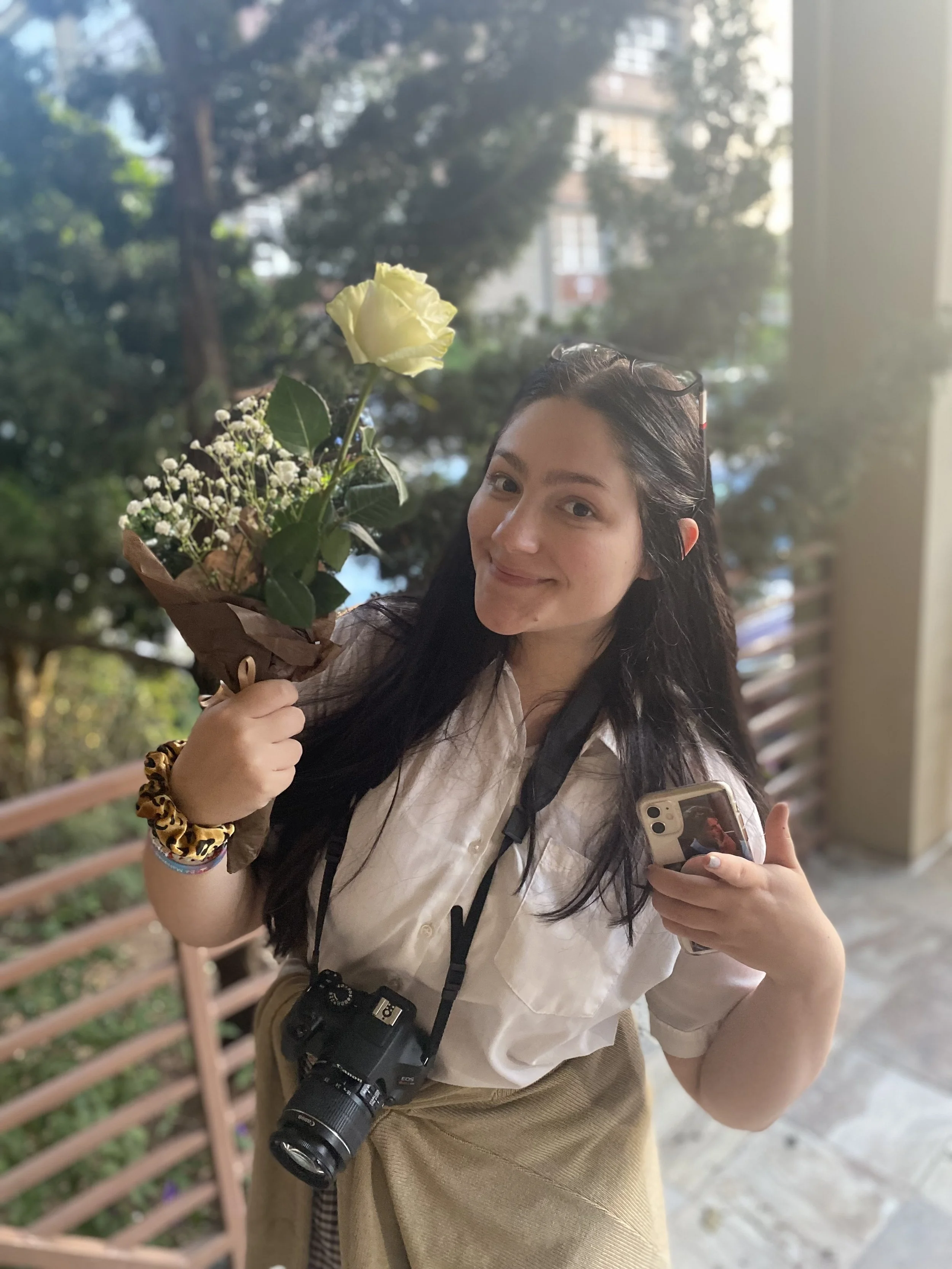 A young woman with long dark hair, smiling, holding a bouquet of white and yellow flowers in one hand and a smartphone in the other. She has a camera hanging around her neck and is standing outdoors near a railing, with trees and buildings in the background.