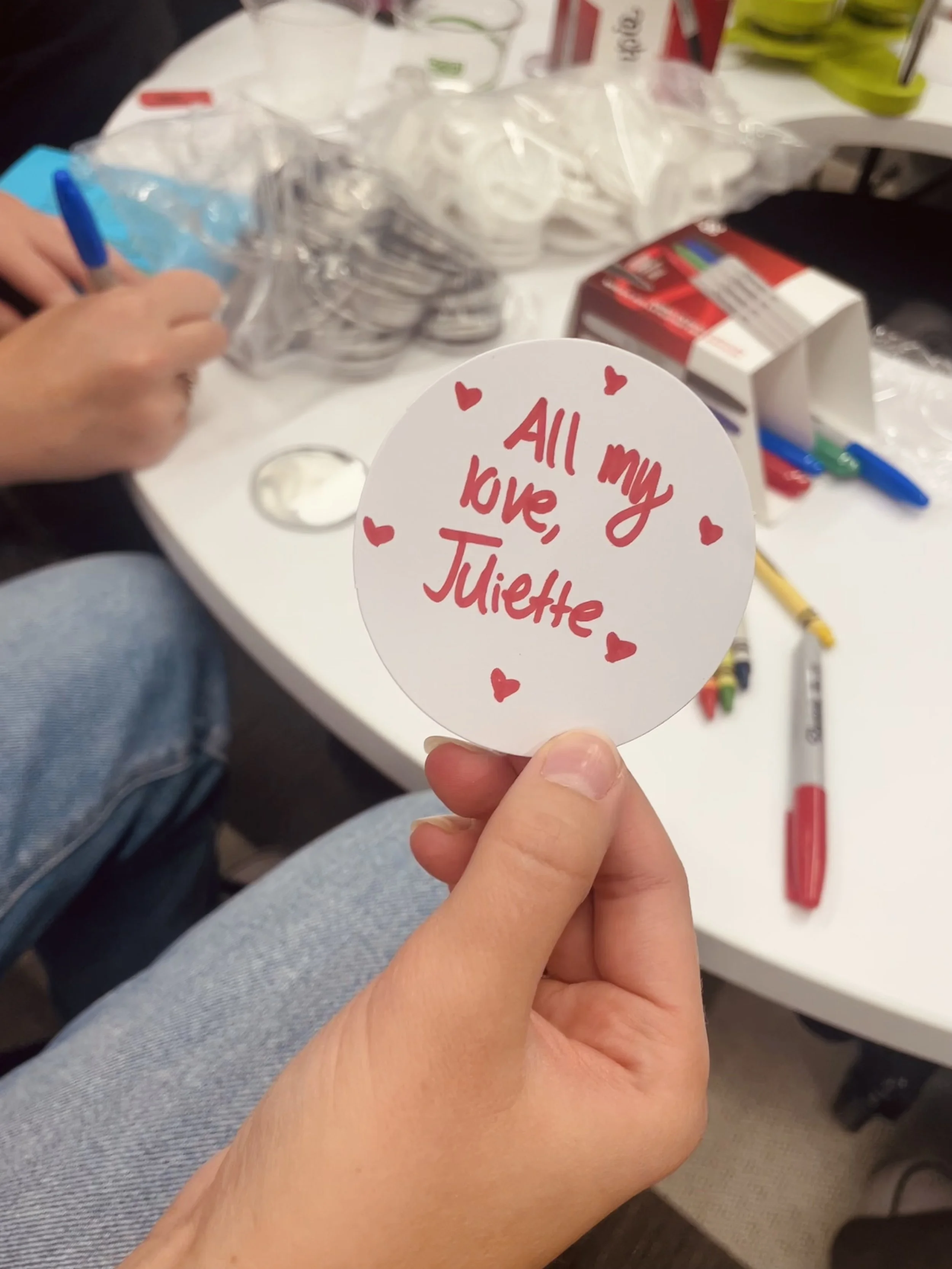 Close-up of a hand holding a round white paper with red handwritten text that says, "All my love, Juliette," surrounded by small red hearts. In the background, there are various markers, packing materials, and a partially visible red and white box on a white table. A person with a blue pen is also visible, sitting at the table.