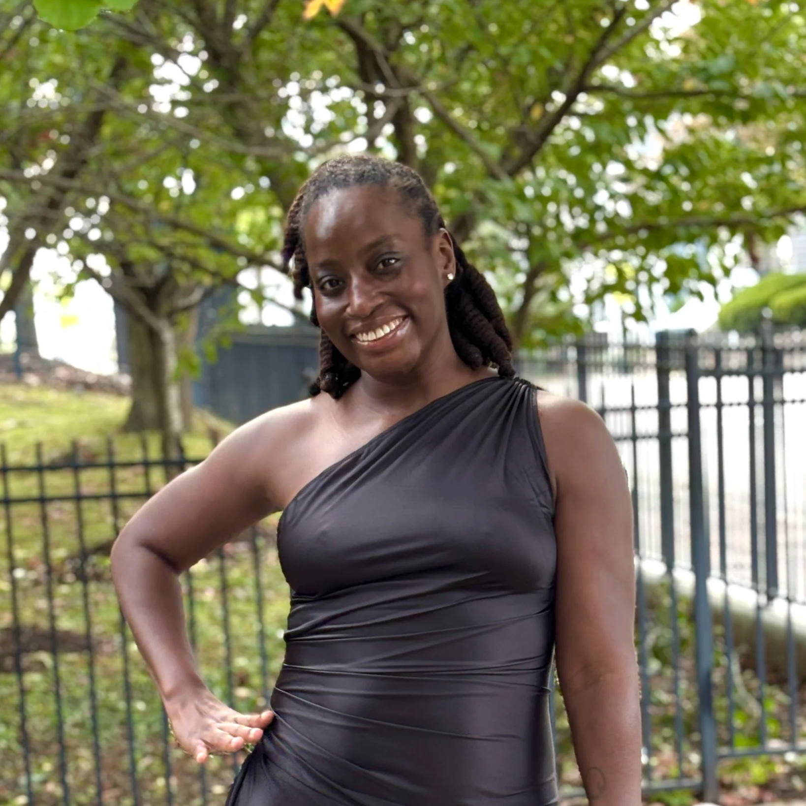 A woman in a sleeveless black dress posing outdoors with trees and a black fence in the background.