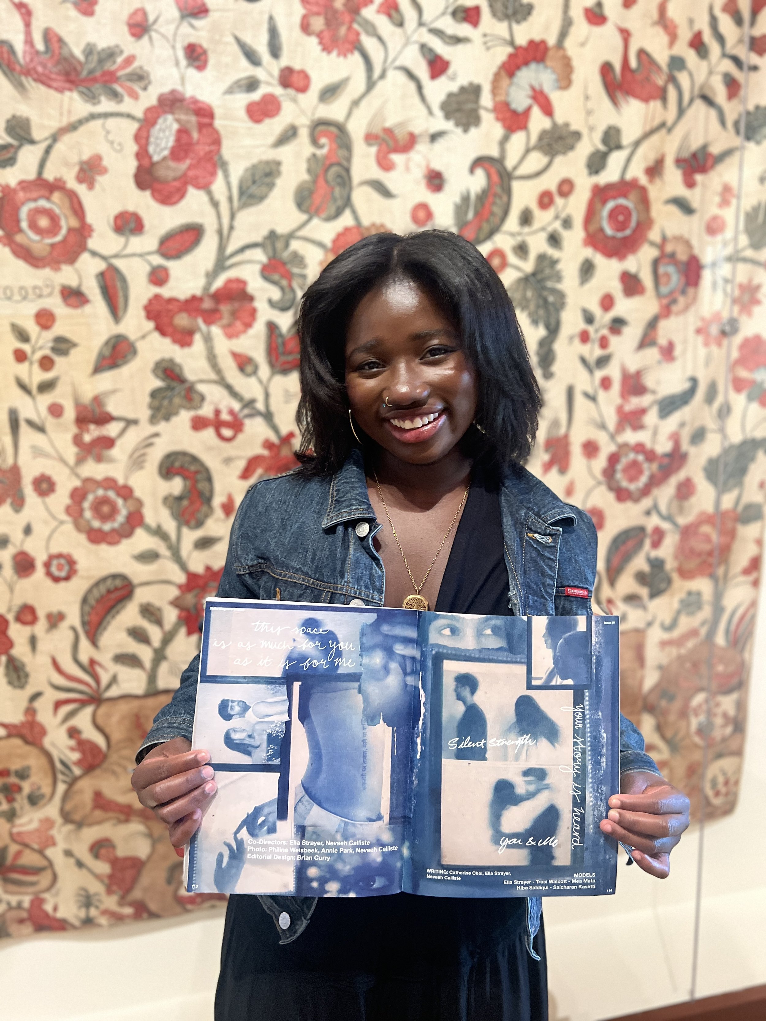 A smiling woman with dark hair and a denim jacket holding an open magazine featuring fashion photos and text, standing in front of a floral-patterned tapestry.