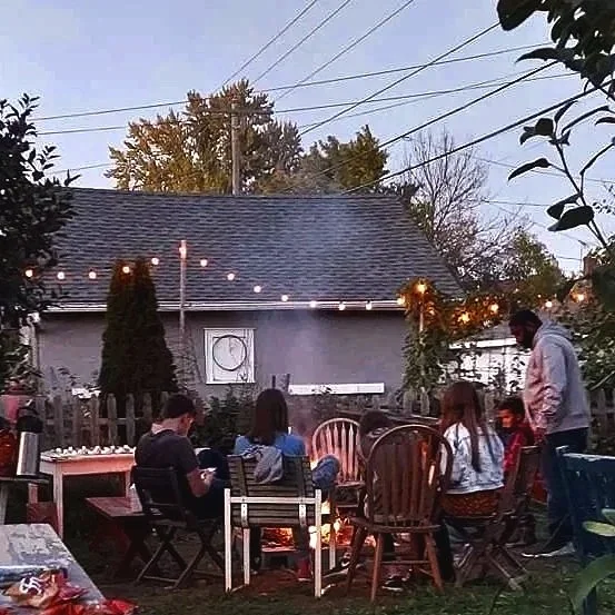 People gathered around a fire pit in a backyard, sitting on chairs and socializing, with string lights hanging overhead and a house in the background.