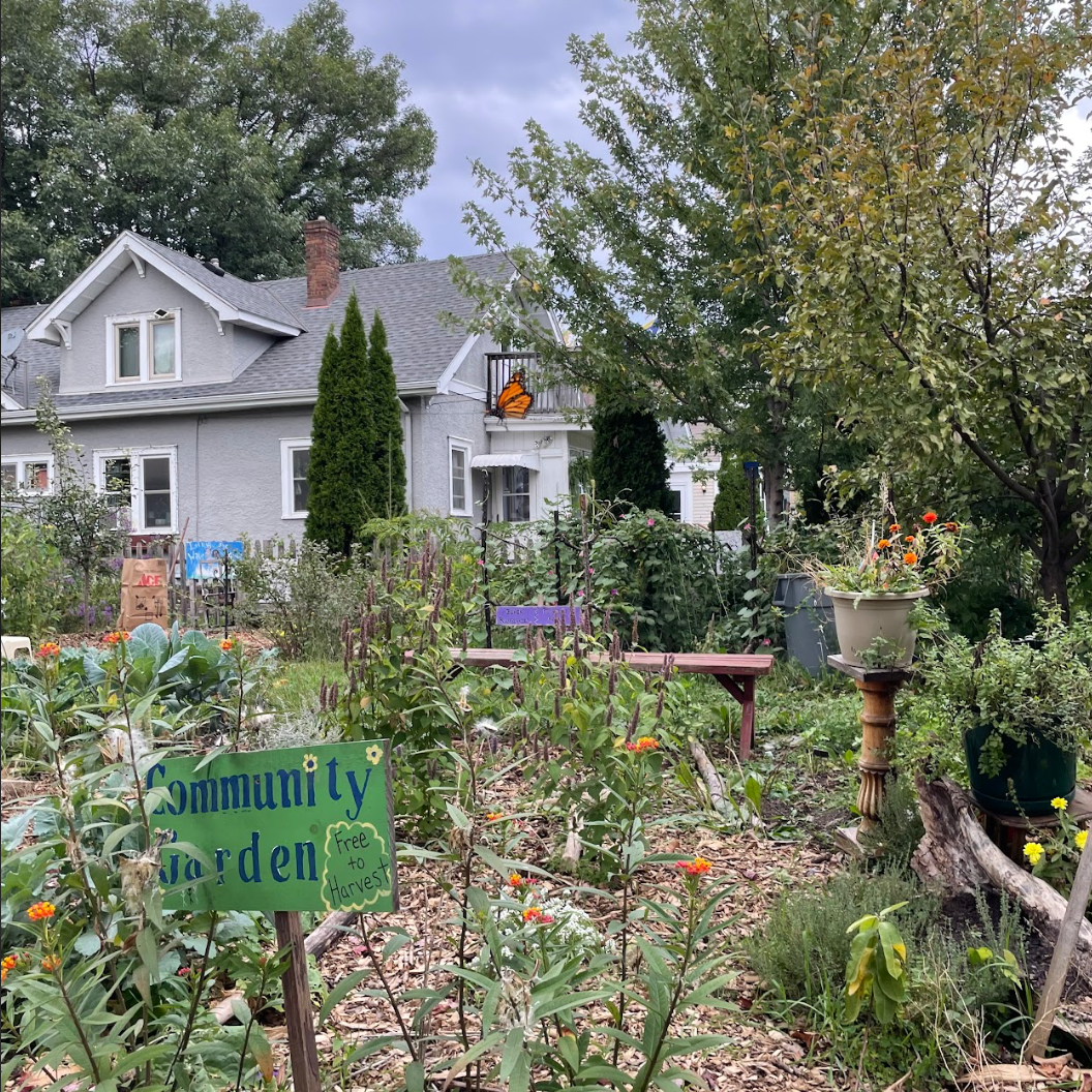 A community garden with various plants and flowers, a sign reading 'Community Garden, Free to Harvest,' and an orange monarch butterfly on a balcony of a light gray house in the background.
