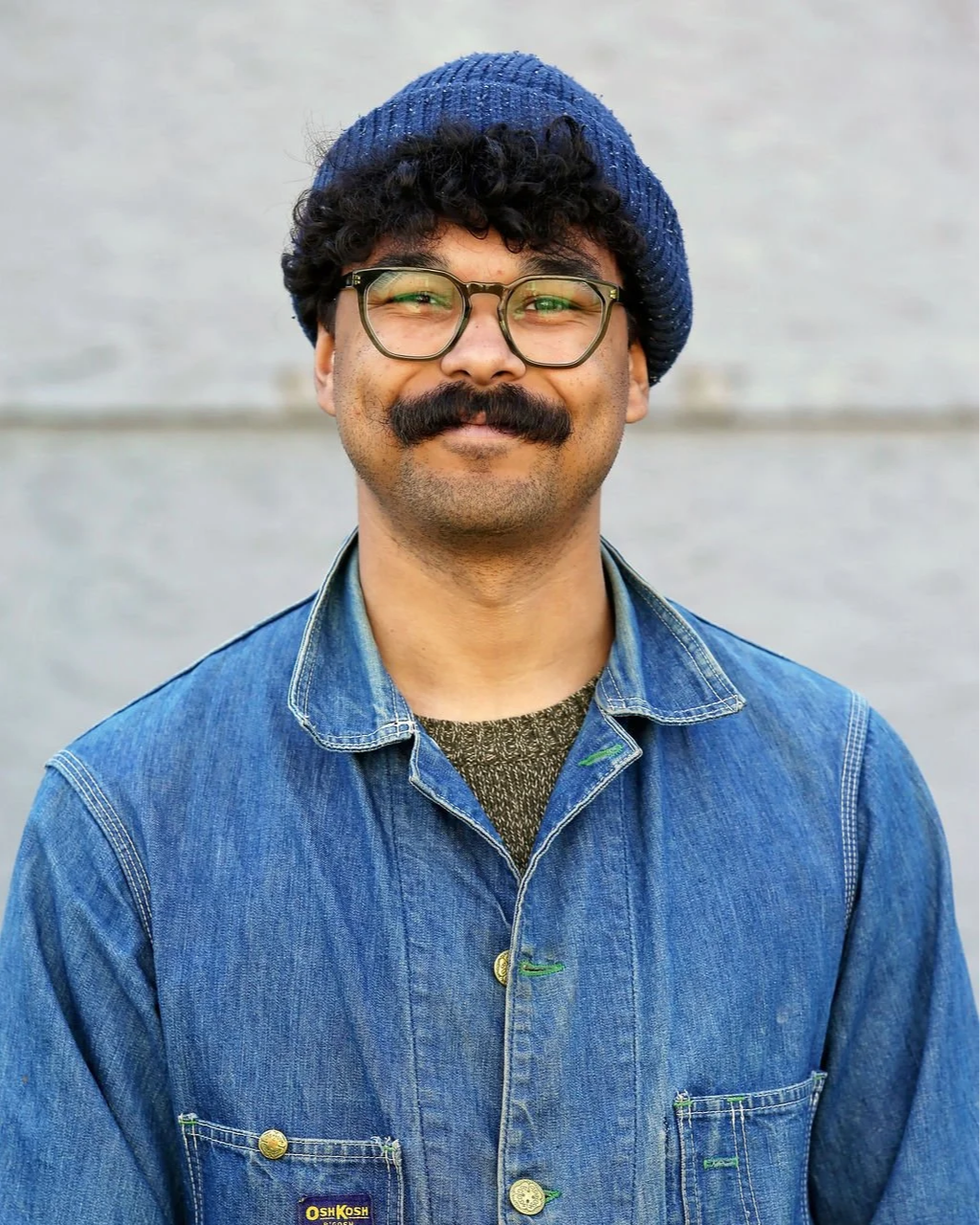 A man with glasses, a mustache, and curly hair, wearing a blue knit hat, denim jacket, and a brown sweater, smiling outdoors against a gray wall.