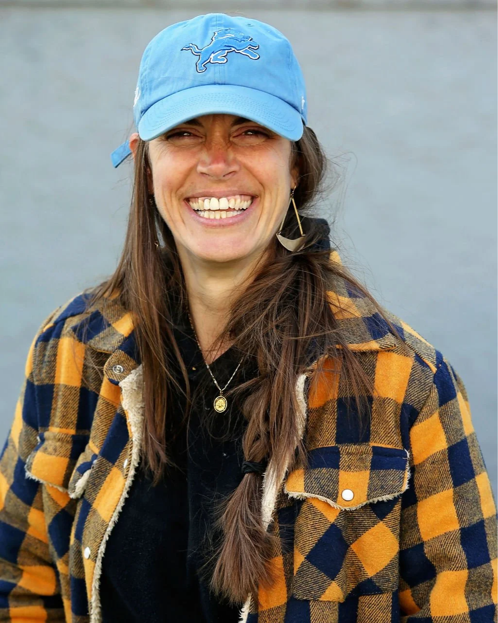 A woman smiling, wearing a blue baseball cap with a Detroit Lions logo and a plaid jacket over a black shirt, standing outdoors.