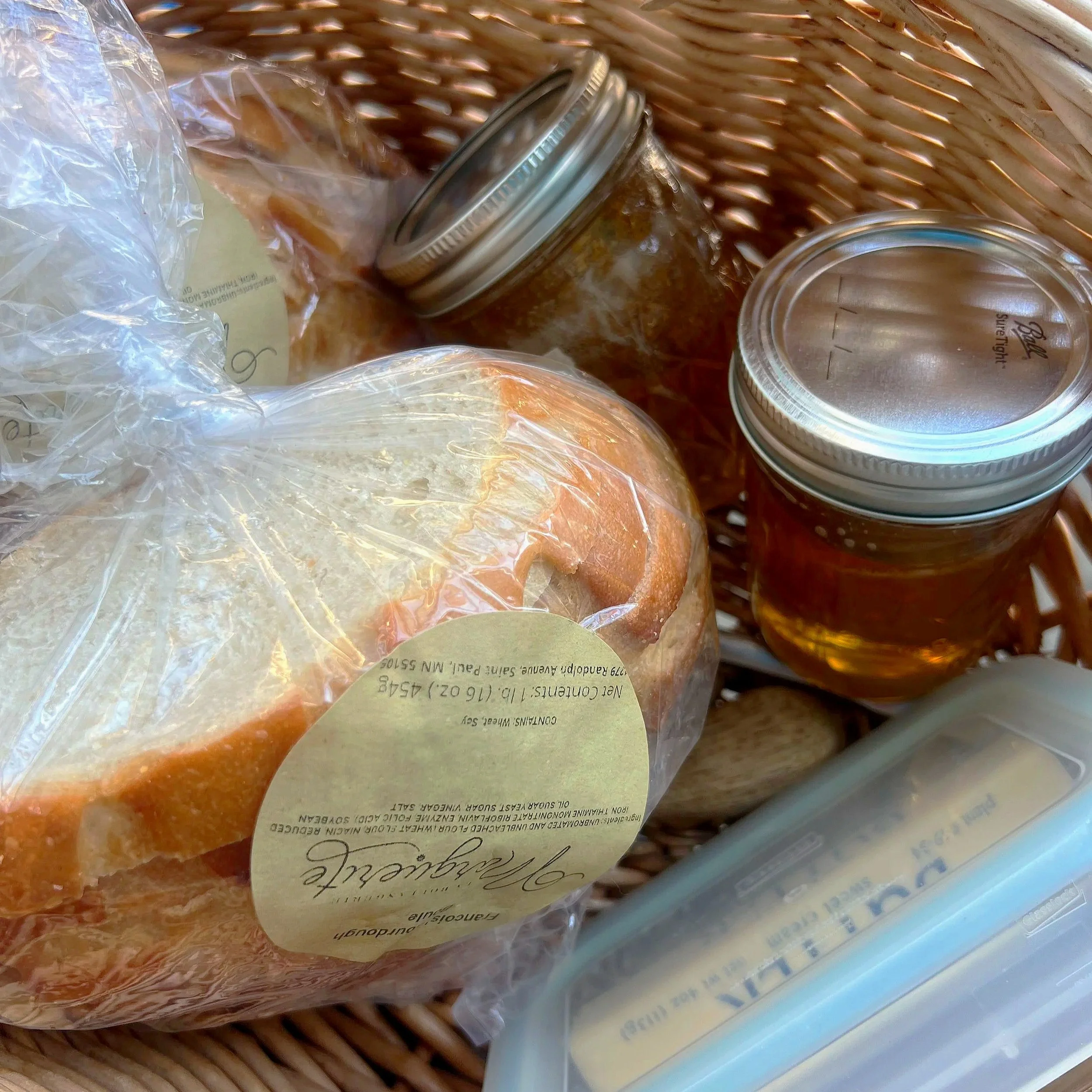 Bread in a plastic bag, jars of honey, and a container of butter in a woven basket.