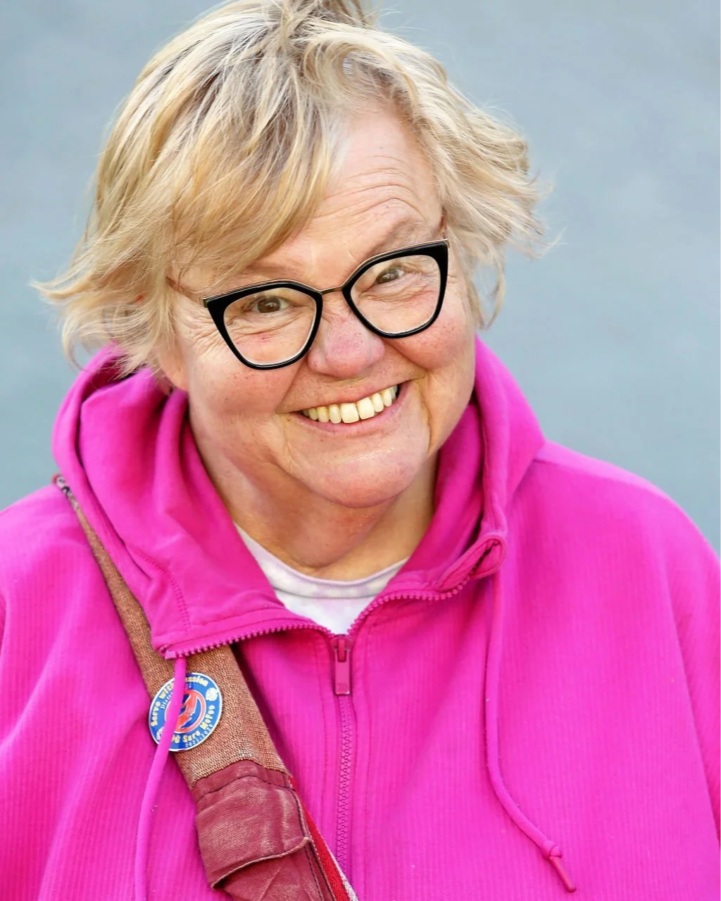 A smiling woman wearing black glasses and a bright pink hoodie with a pin on her strap.