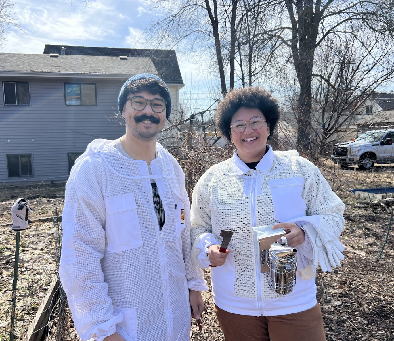 A man and woman smiling outdoors in a yard with bare trees and a house in the background. The man has glasses, a mustache, and a beard, and is wearing a white jacket and a blue knit hat. The woman has glasses and an afro hairstyle, and is wearing a white jacket, holding gardening gloves, a small metal watering can, and a gardening tool.