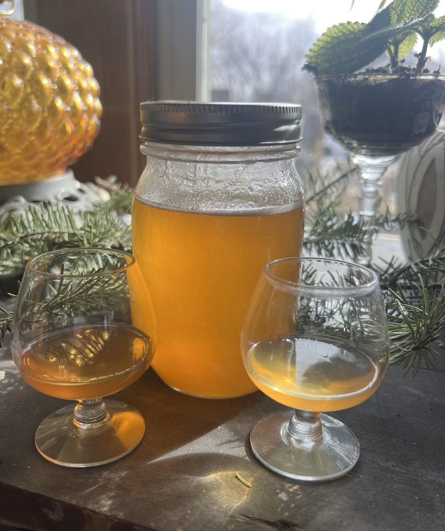 A jar of honey and two snifter glasses with a golden liquor sit on a table decorated with evergreen sprigs. A potted plant is visible in the background.