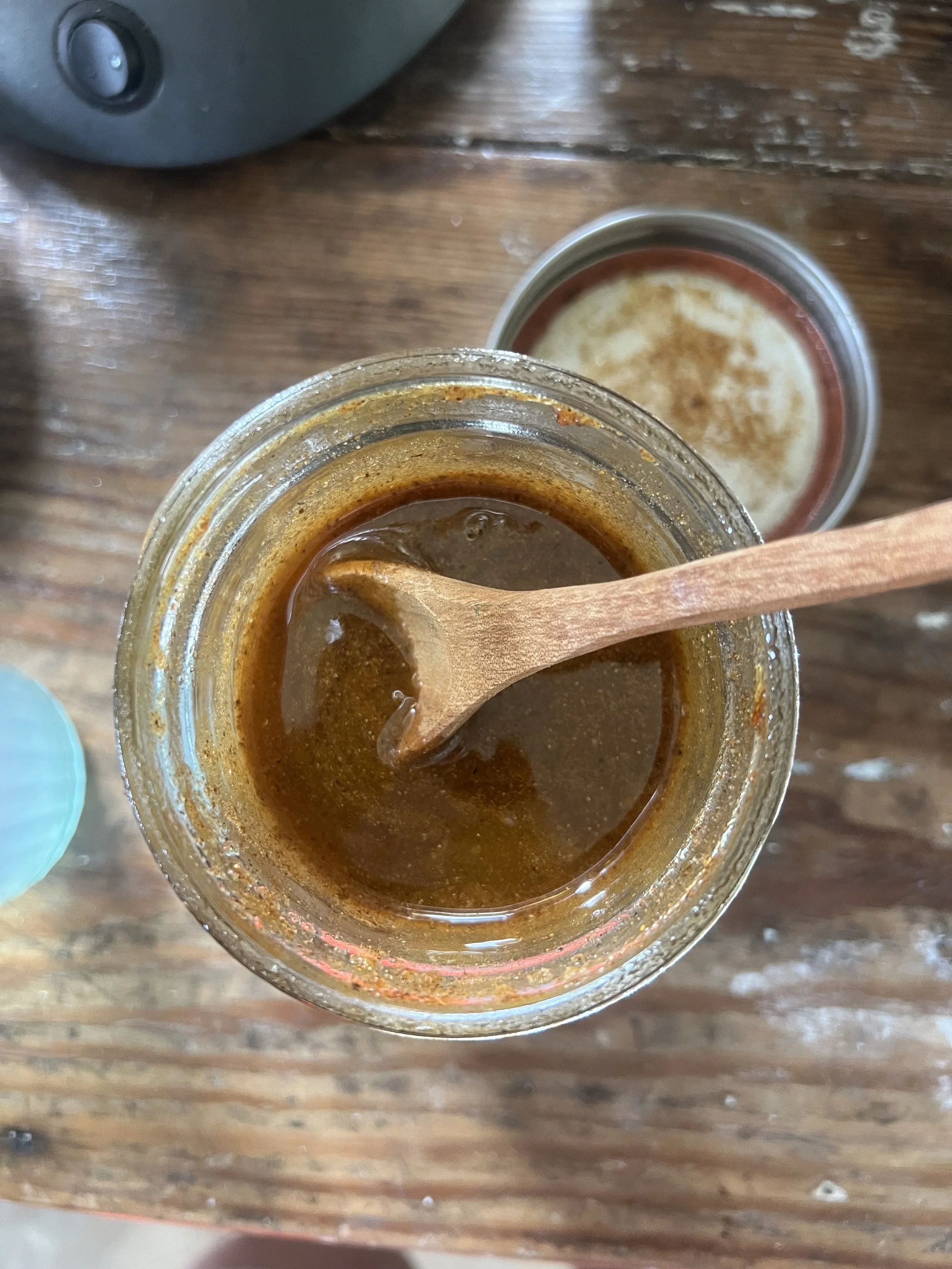 A jar of thick, dark brown honey with a wooden spoon inside it, and a glass of milk with cinnamon on top on a wooden table.