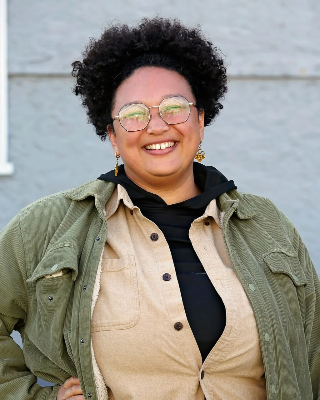 A woman with curly black hair, glasses, and earrings, smiling outdoors against a gray wall, dressed in layered casual clothing including a green jacket and beige shirt.