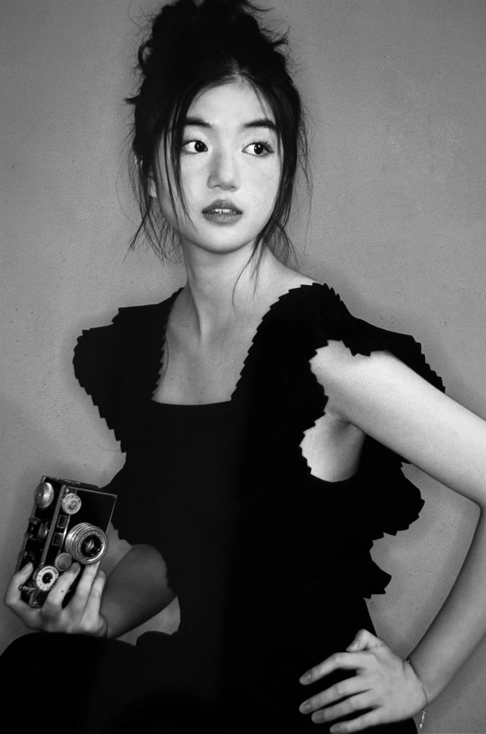 A young woman with dark hair styled in a messy bun, wearing a black dress with textured sleeves, holding a vintage camera, standing against a plain background.